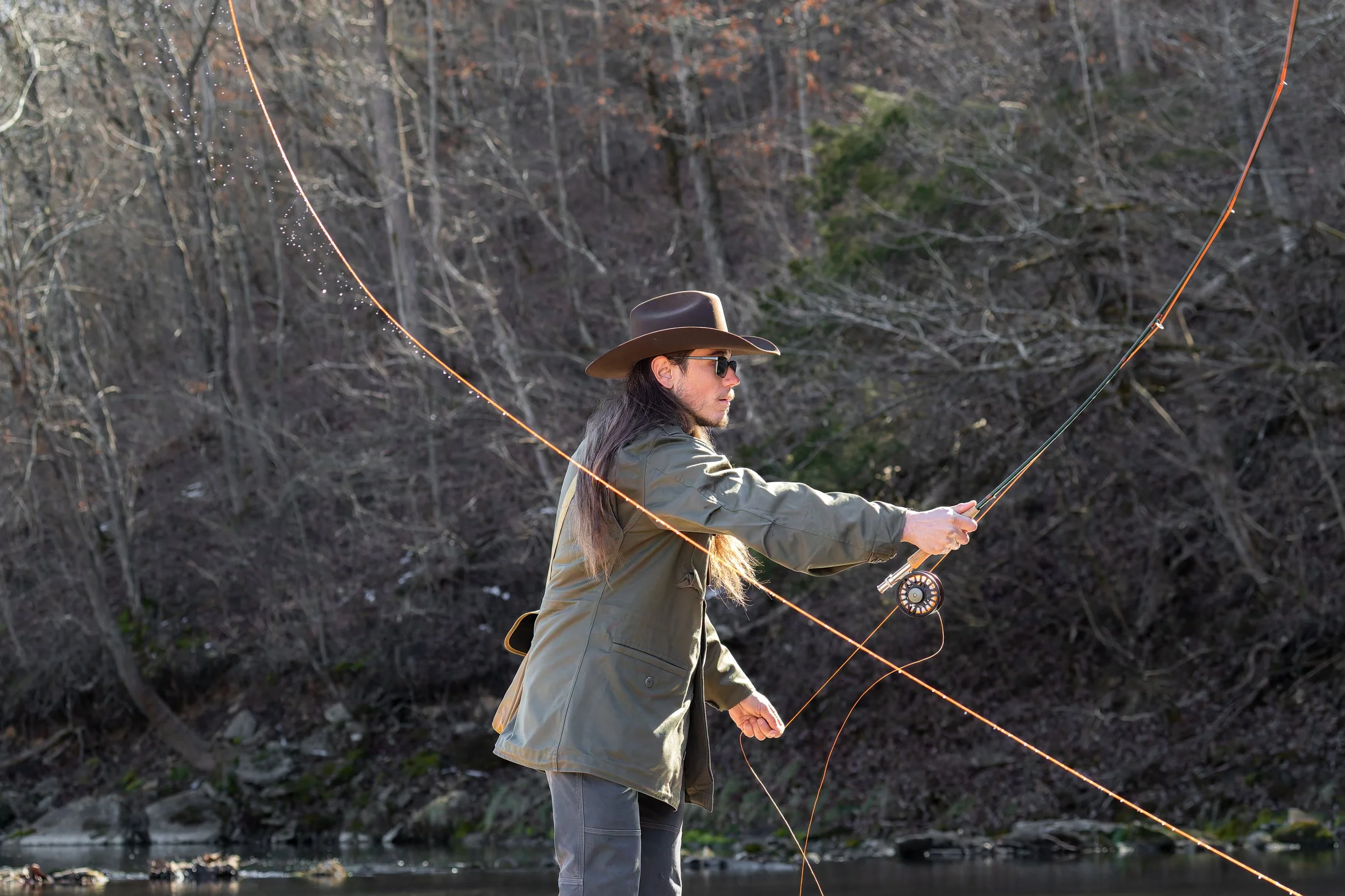 A man fishing in a river, wearing a wide-brimmed hat, sunglasses, and a green jacket, casting his fishing line with a rod in a natural outdoor setting with leafless trees and rocks in the background.