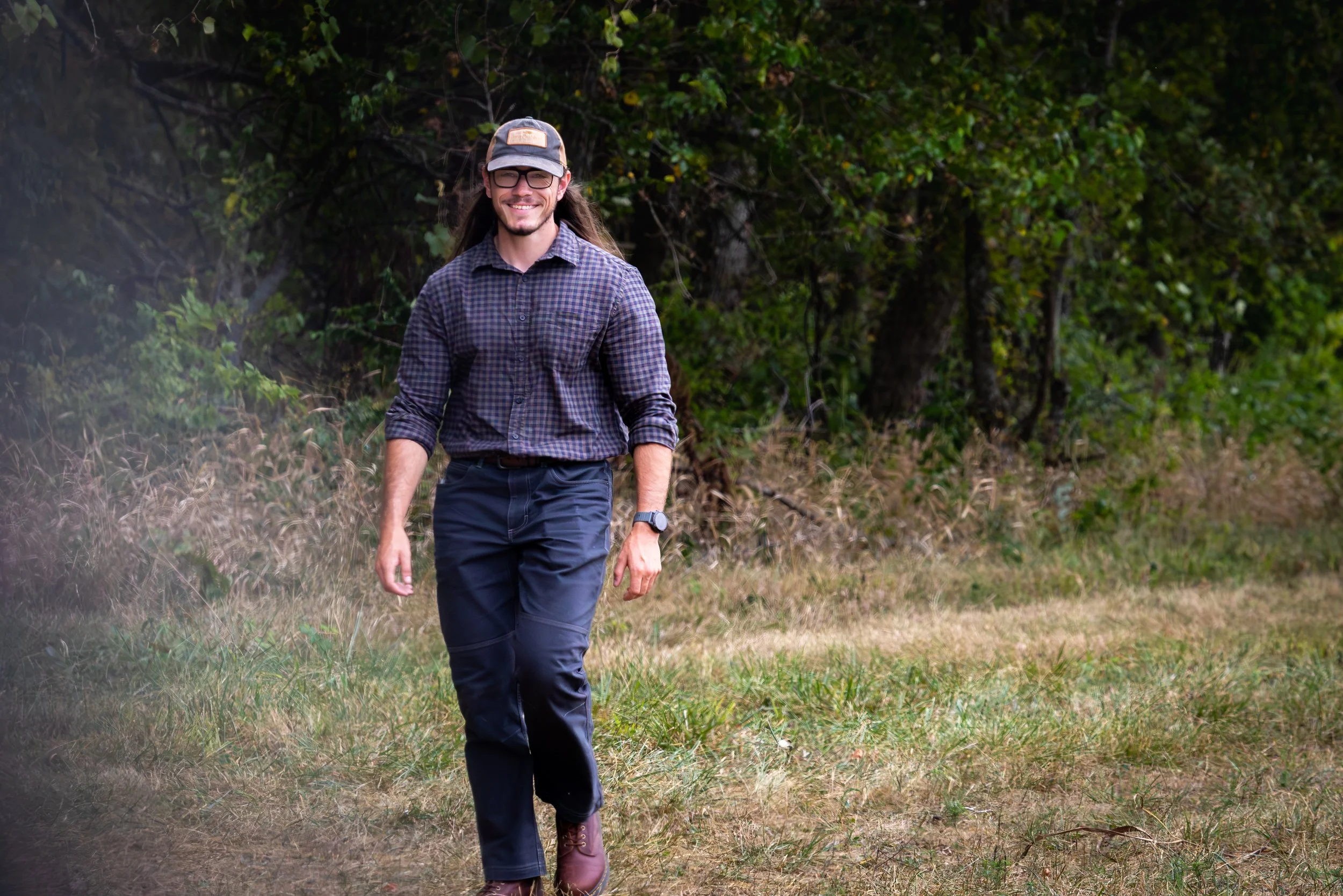 A man with long hair, glasses, and a beard walking outdoors in a grassy area with trees in the background. He is smiling and wearing a cap, a checkered shirt, dark pants, and boots.