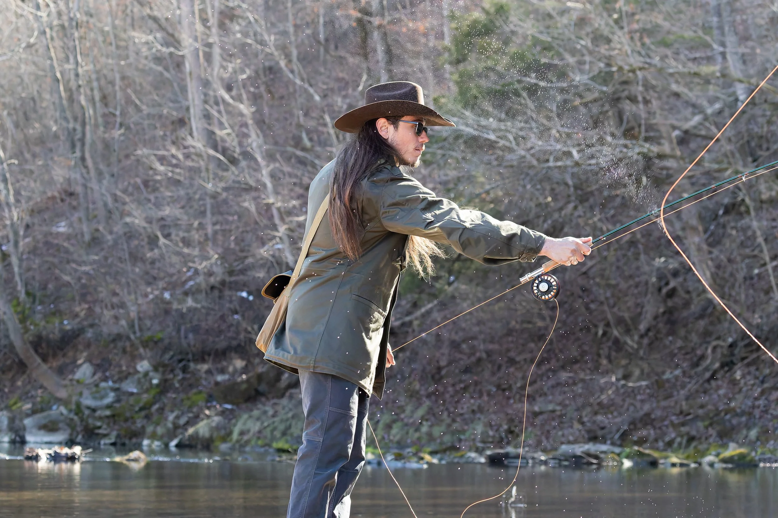 Person fishing in a river, wearing a hat and sunglasses, casting a line with a fishing rod.