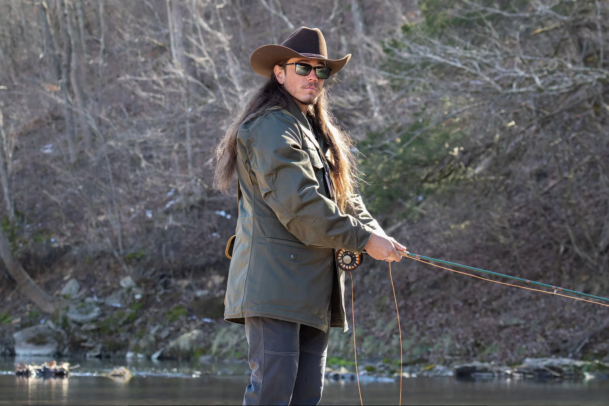 A man with long hair, wearing sunglasses, a wide-brimmed hat, a green jacket, and dark pants, fishing in a river surrounded by trees in early spring or fall.