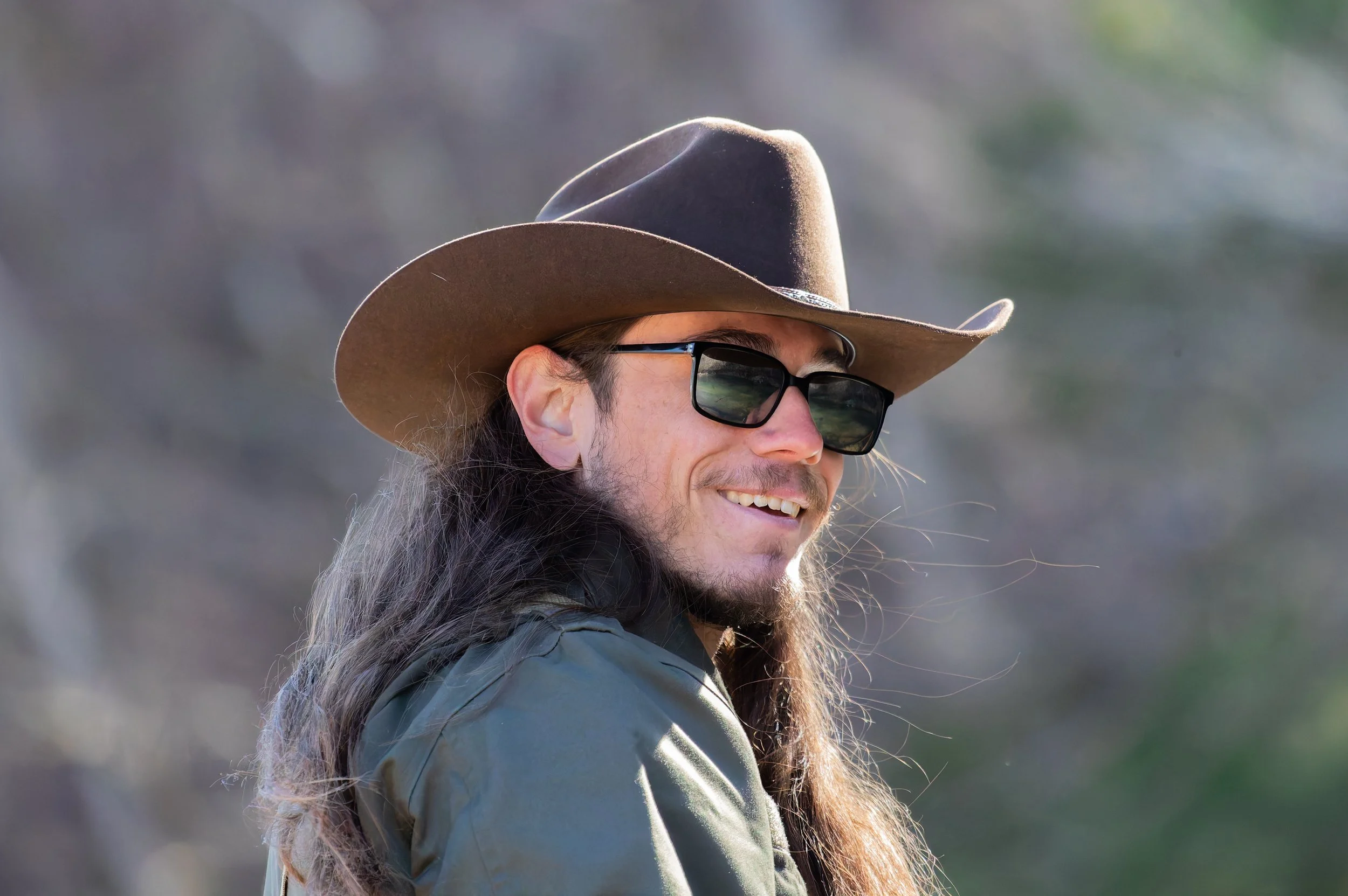 Man wearing a brown cowboy hat, black sunglasses, and a green jacket, smiling outdoors during daytime.