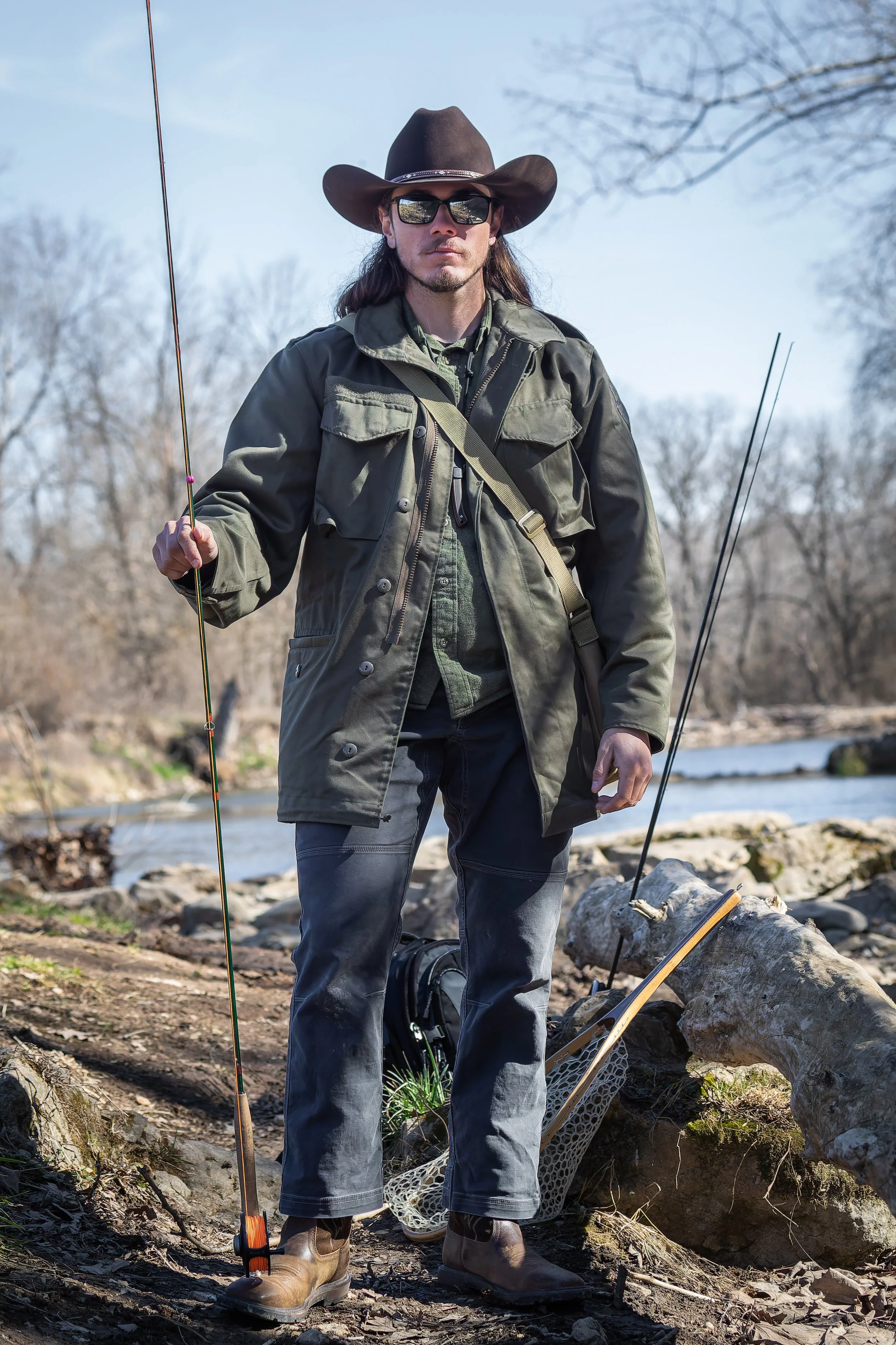 A man in outdoor fishing gear standing by a river with fishing rods, wearing sunglasses, a wide-brimmed hat, a green jacket, dark pants, and brown boots.