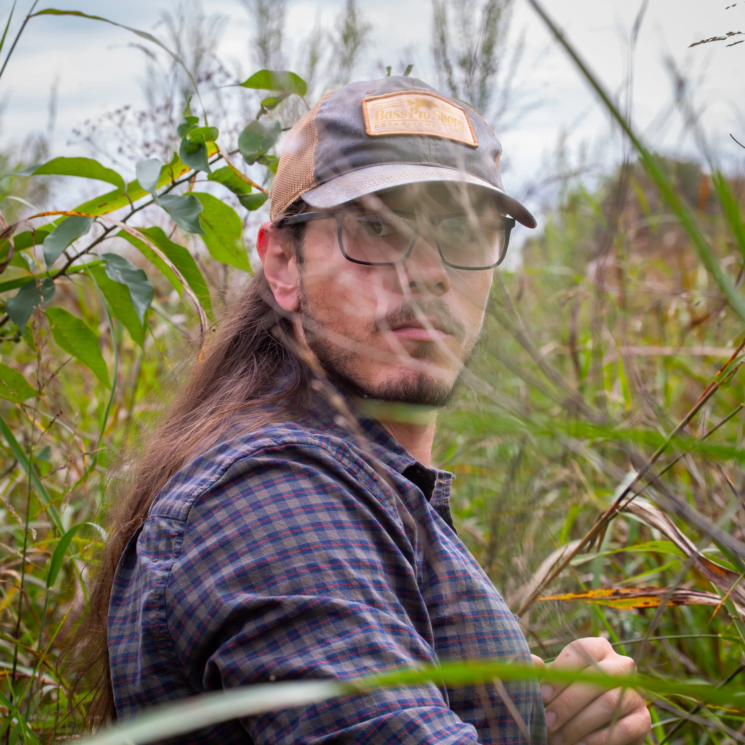 A man with long hair, glasses, and a beard wearing a cap and plaid shirt is standing among tall grass and plants in an outdoor setting, looking directly at the camera.