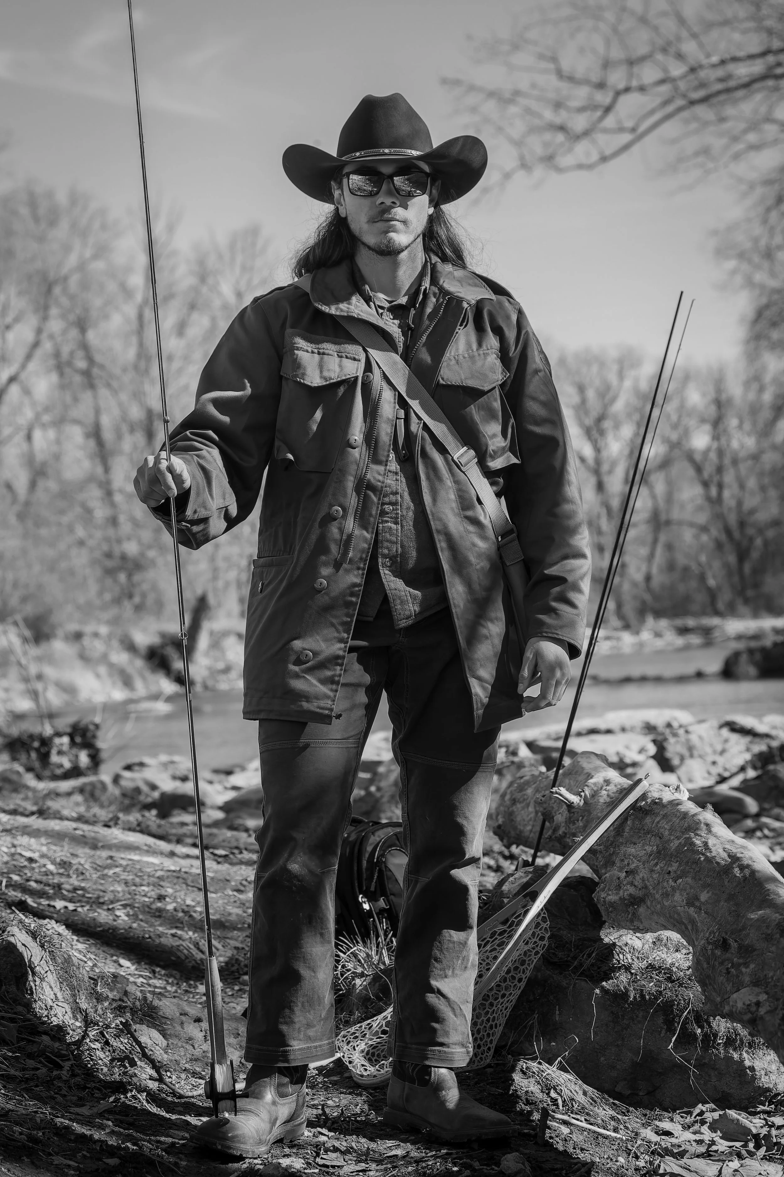 A man with long hair wearing a cowboy hat, sunglasses, a jacket, and jeans, standing outdoors near a river with trees in the background. He is holding a fishing pole and has a fishing net on the ground beside him.