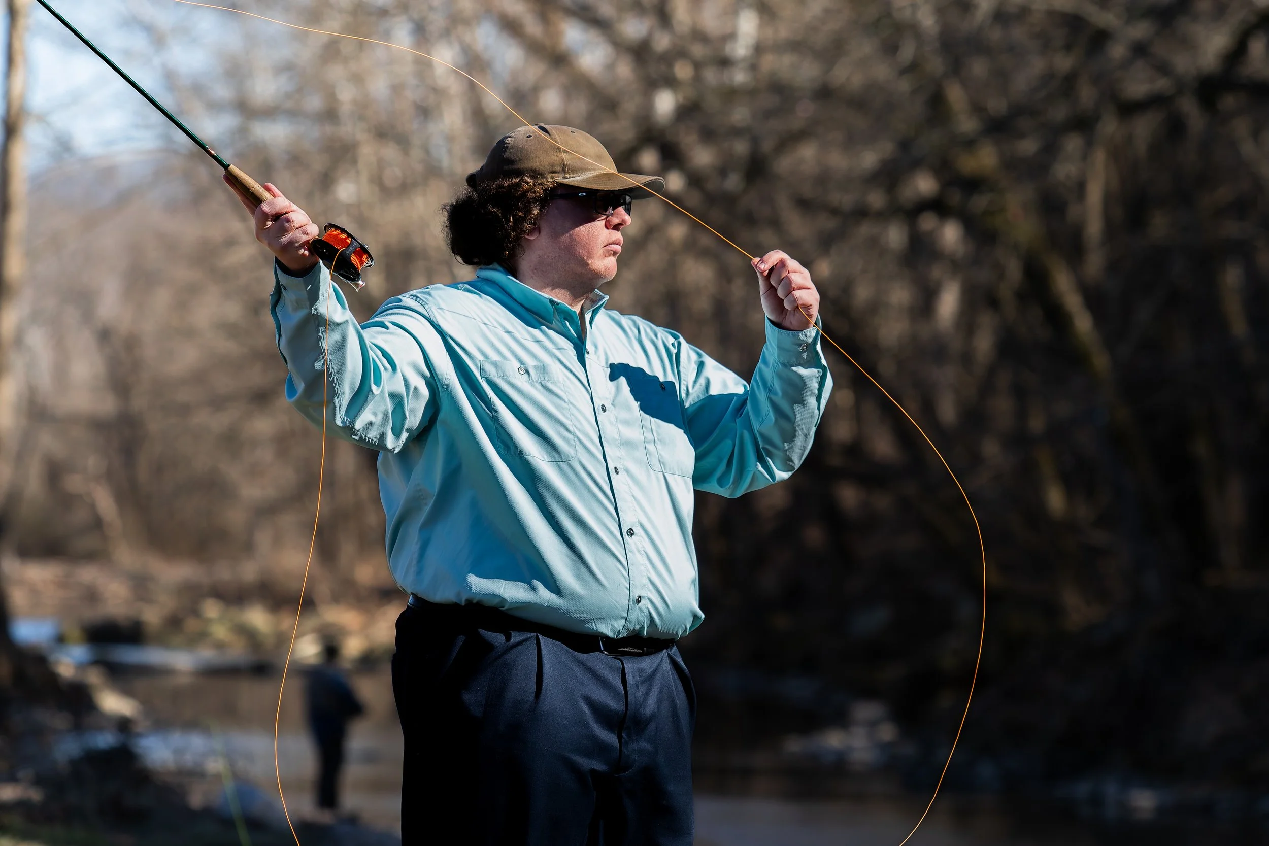 A man in a light blue shirt, sunglasses, and a brown cap is fishing with a fishing rod near a river, with bare trees in the background.