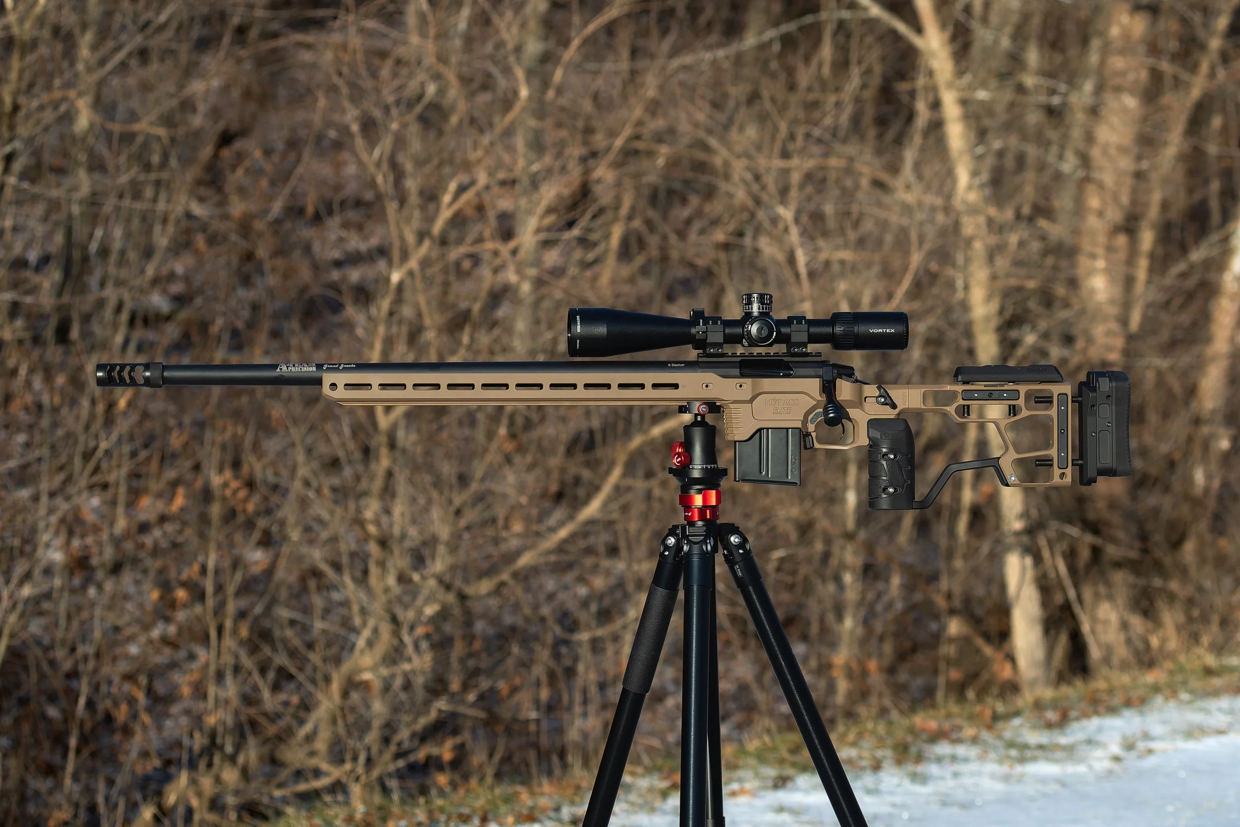 A sniper rifle with a scope mounted on a bipod stand outdoors with bare trees in the background.