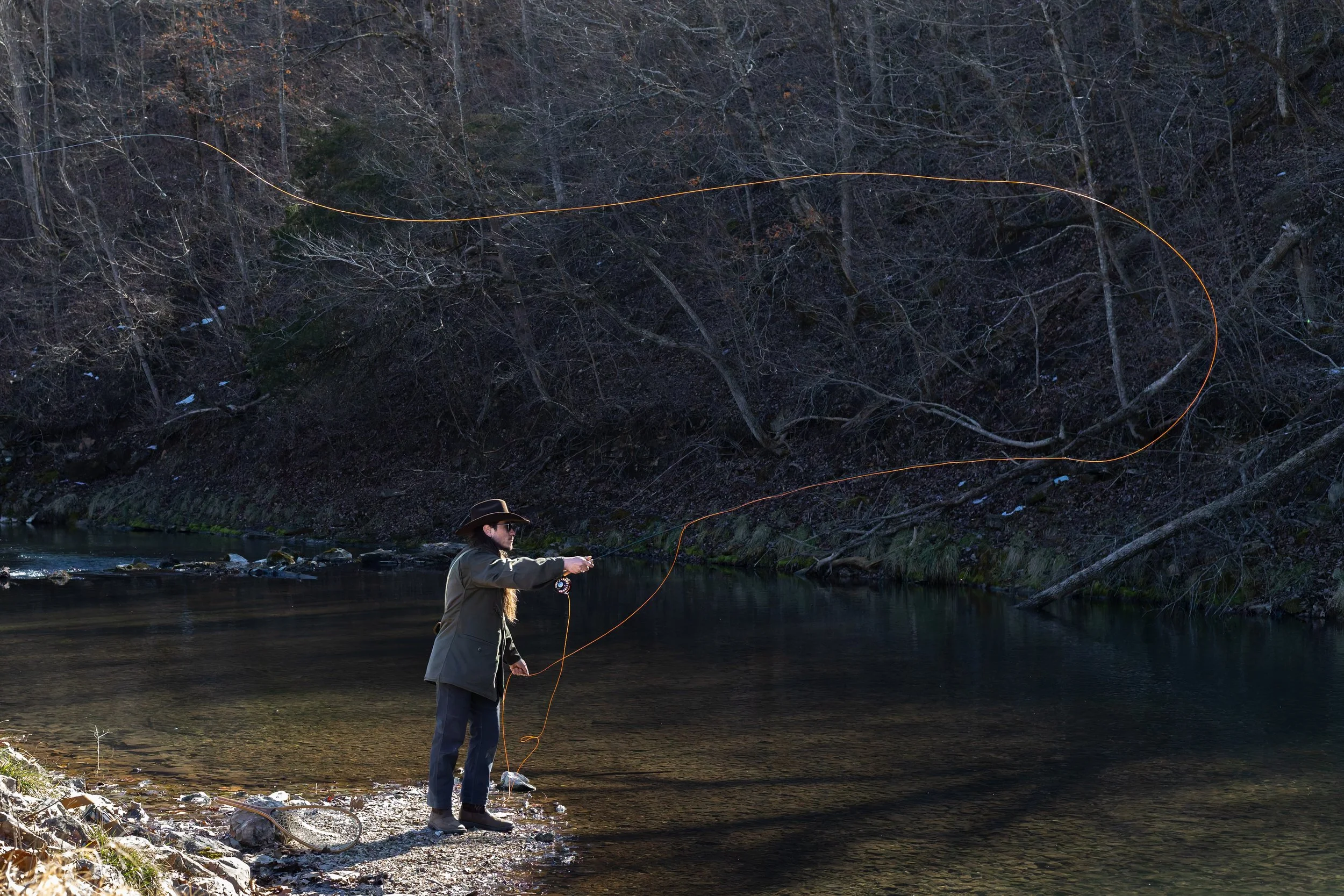 A woman fishing in a river with a rod, standing on the riverbank, surrounded by a wooded area with bare trees.