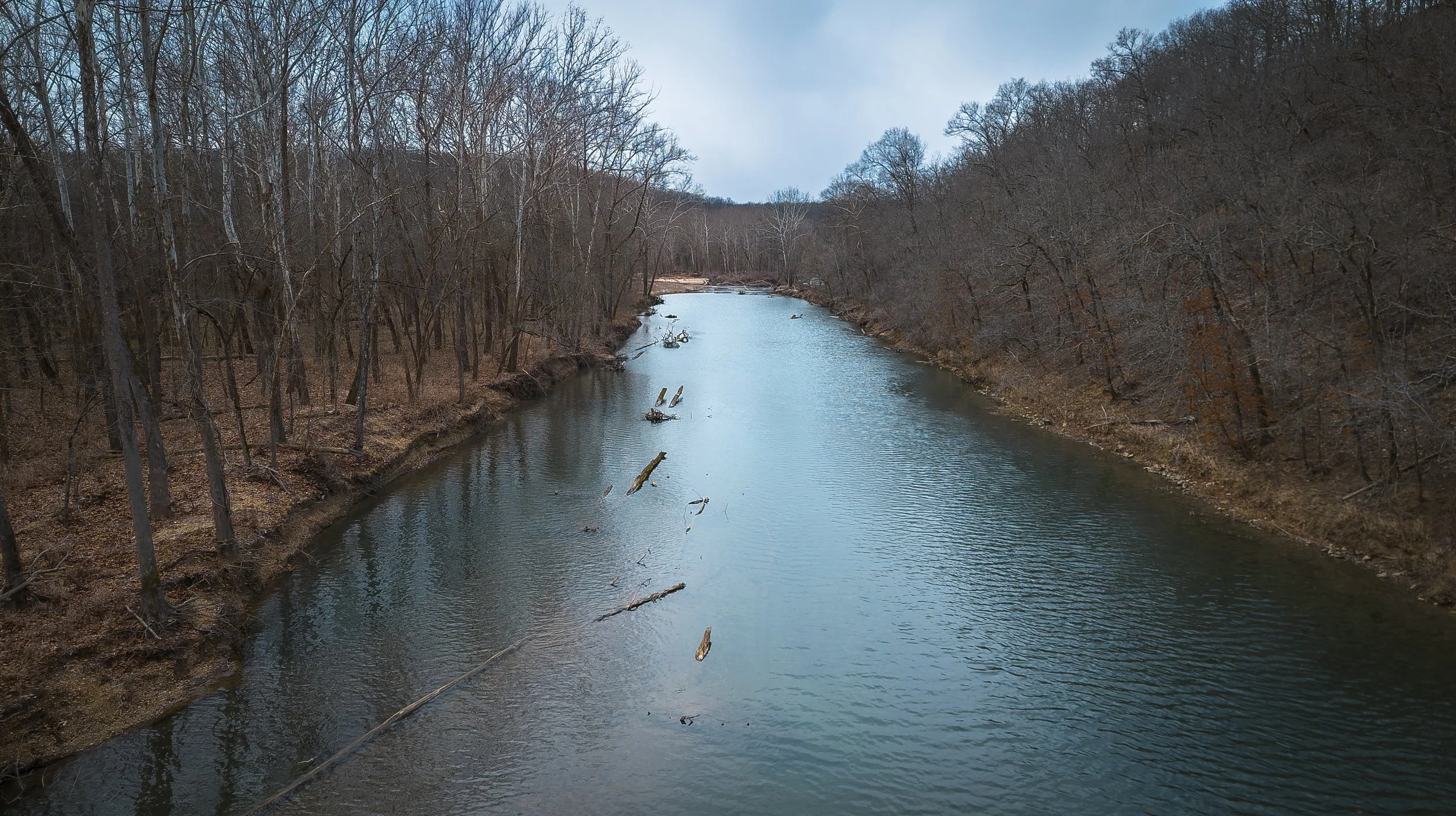 A river flowing through a wooded area with leafless trees on both sides, some fallen branches in the water, and a cloudy sky overhead.