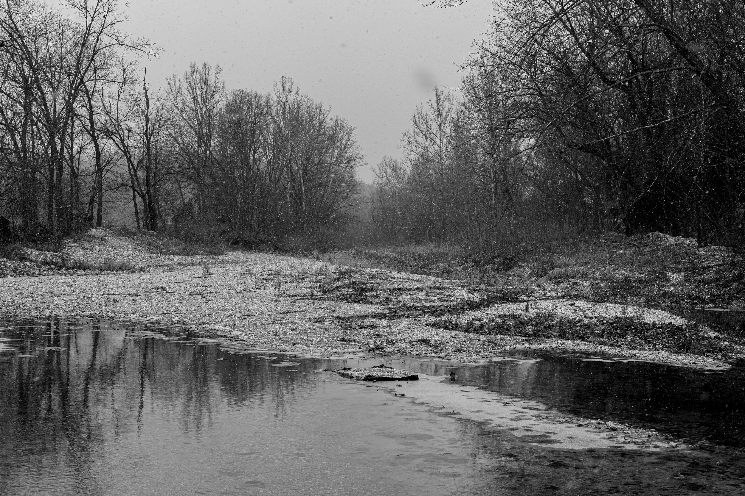 A black and white photo of a river with ice and snow along its banks, surrounded by leafless trees in winter.