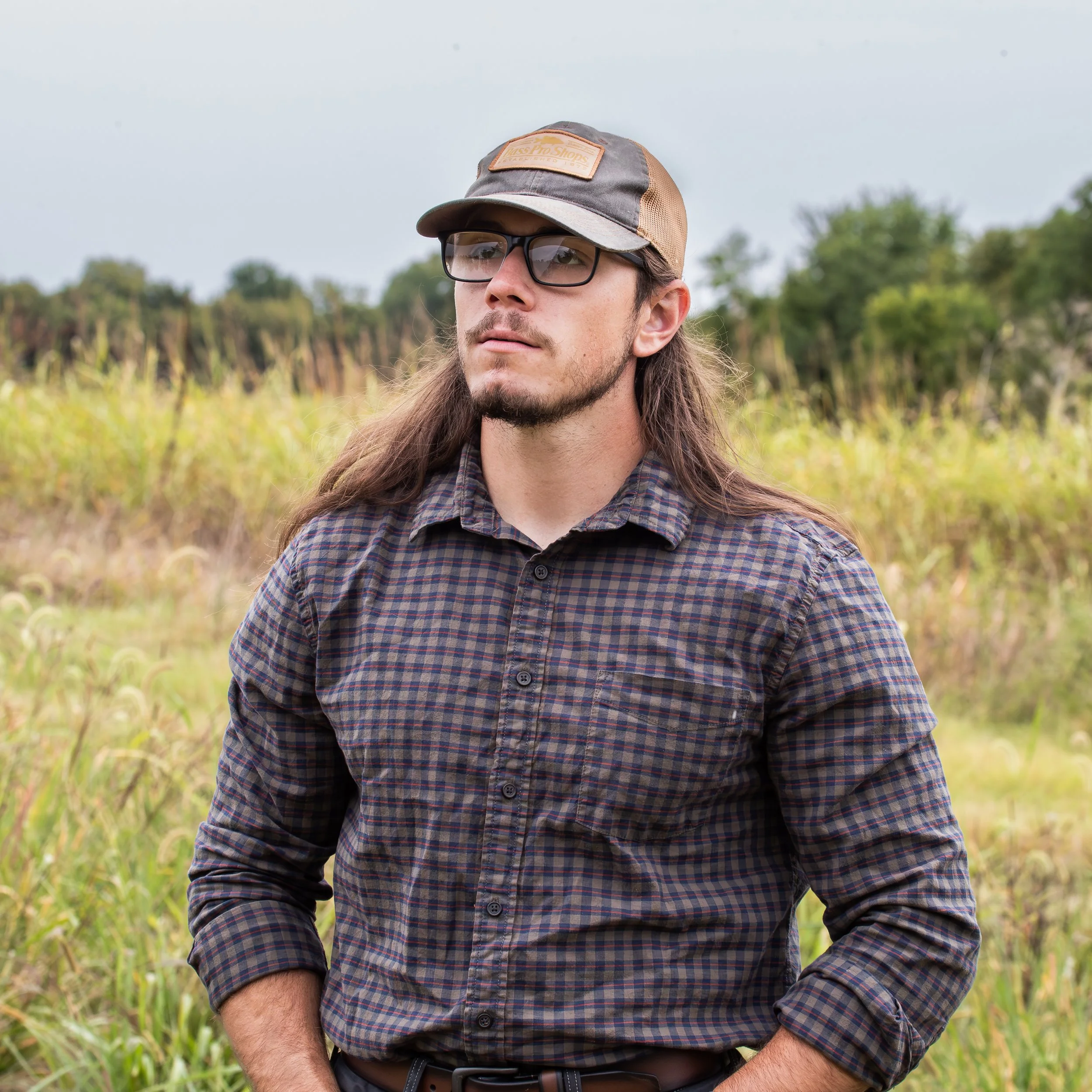 Young man with long hair, beard, glasses, wearing a checked shirt and a cap, standing outdoors in a field with tall grass and trees in the background.