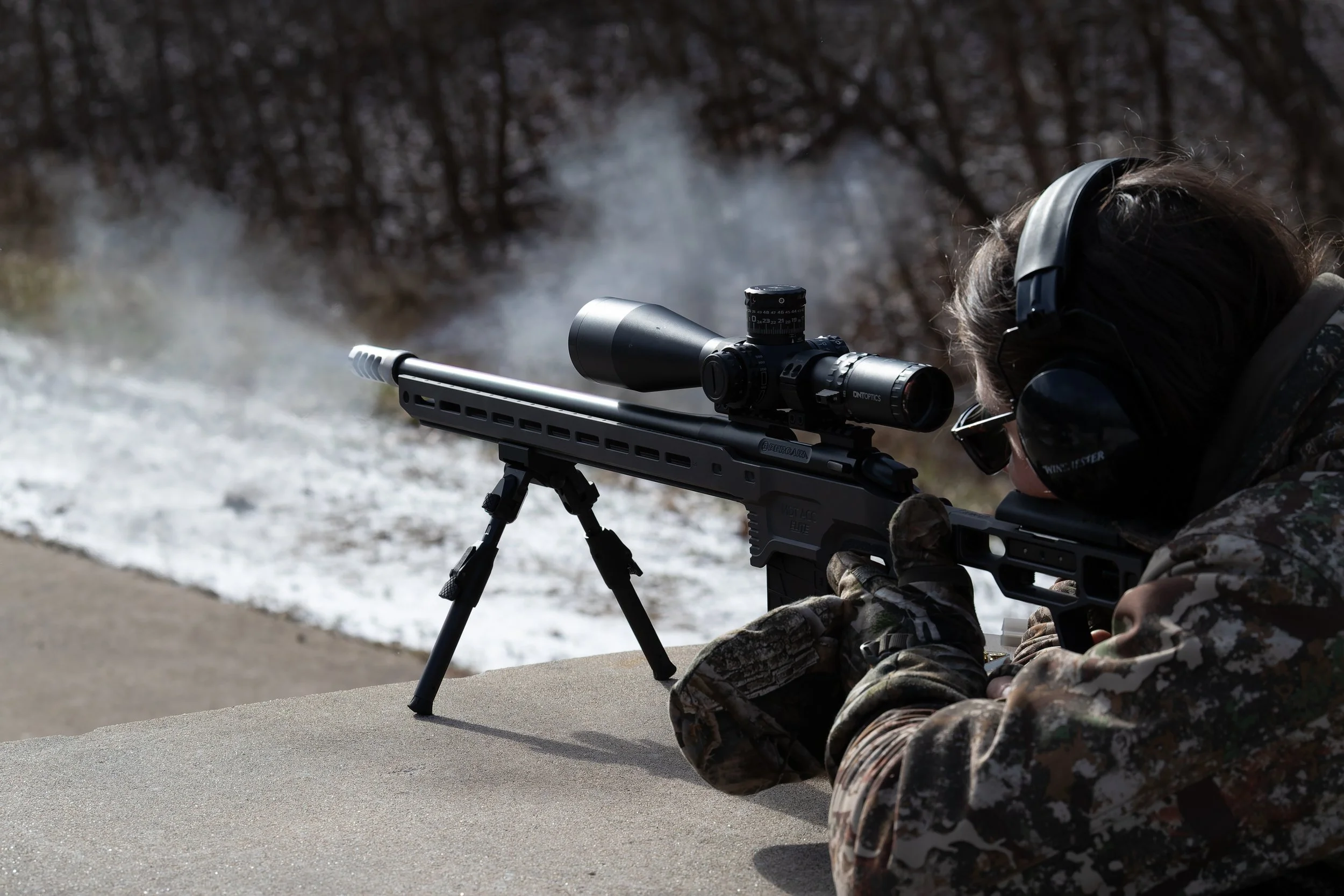 Person in camouflage clothing aiming a sniper rifle with a scope on a bipod, outdoors in a winter setting.