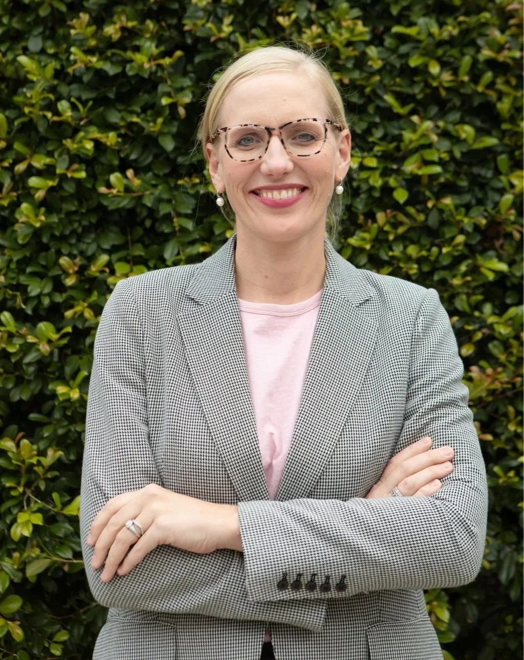 A woman with blonde hair and glasses smiling with arms crossed, standing outdoors in front of a leafy green hedge, wearing a gray blazer, pink shirt, and pearl earrings.