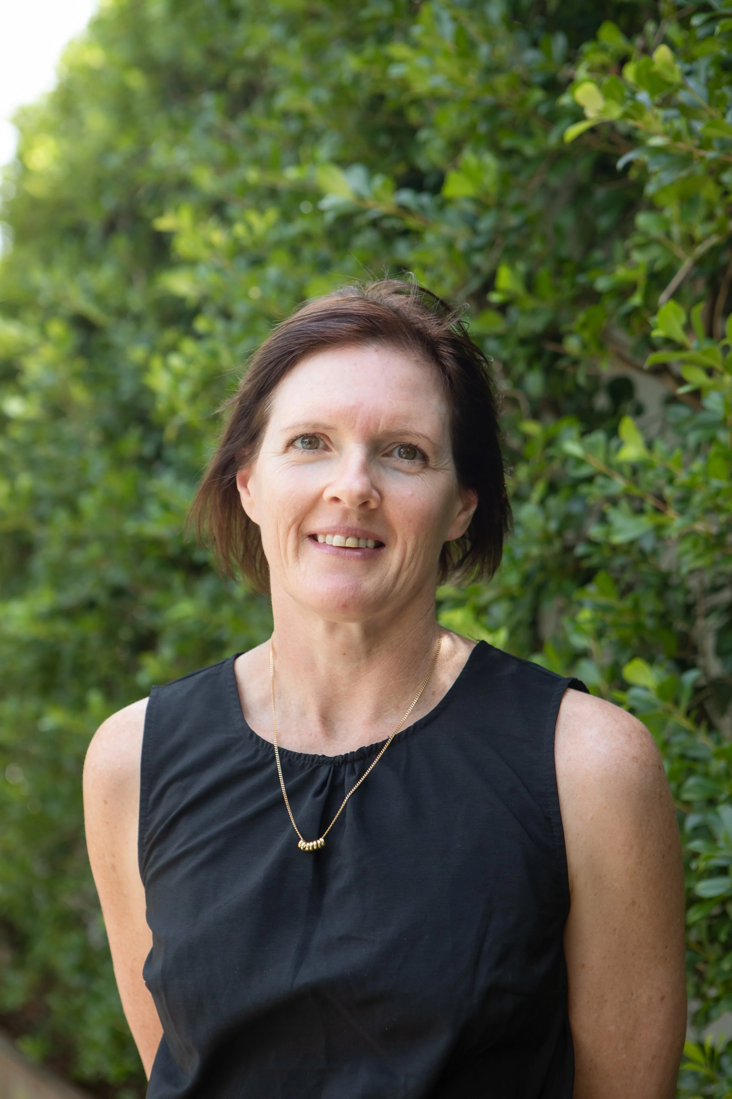 A woman with short brown hair wearing a black sleeveless top and a gold necklace, standing outdoors in front of green foliage.