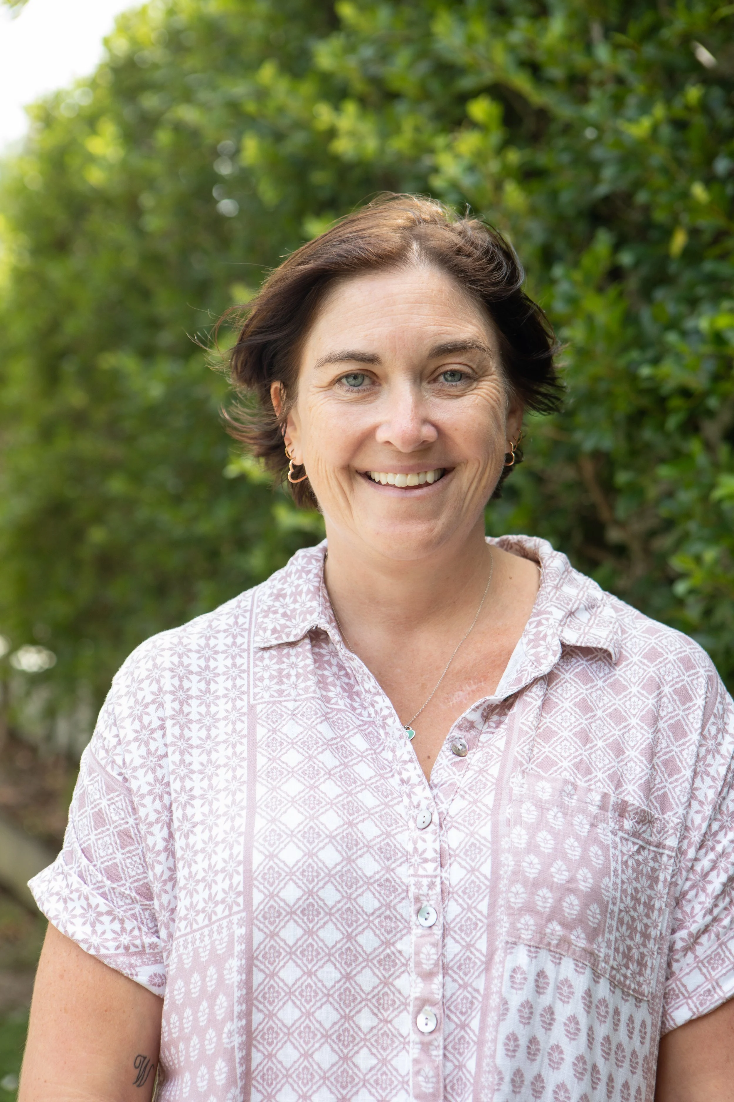 A woman with short brown hair, blue eyes, and a light pink patterned shirt standing outdoors with green foliage in the background.
