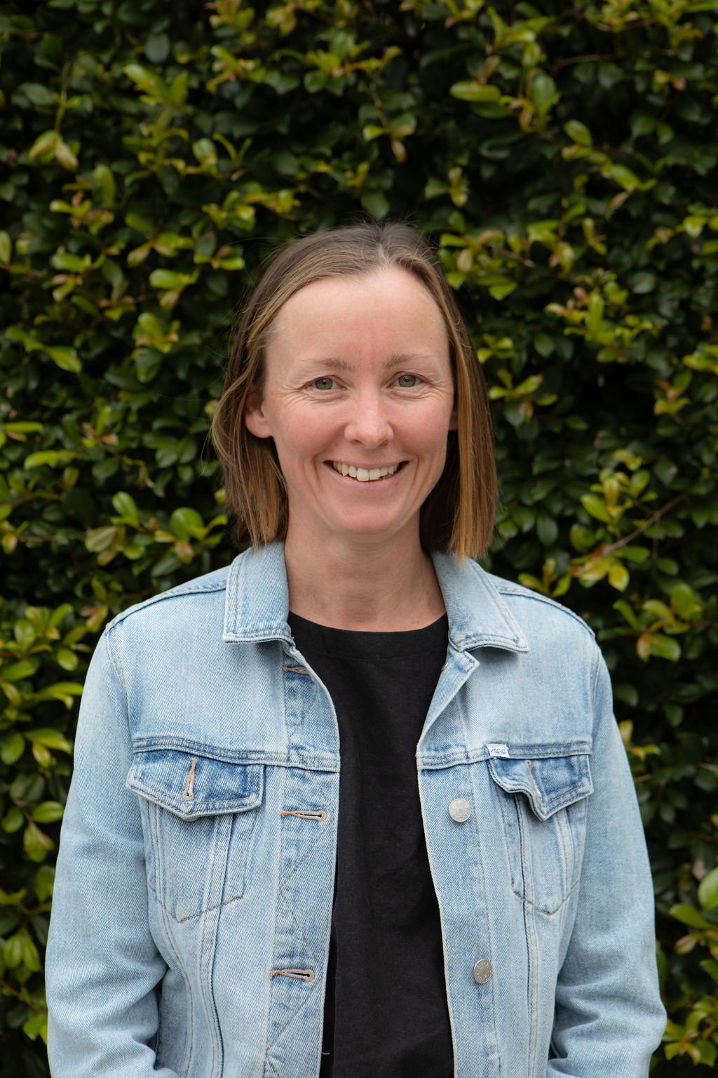 A woman with short brown hair, smiling, wearing a light blue denim jacket over a black shirt, stands in front of green leafy bushes.