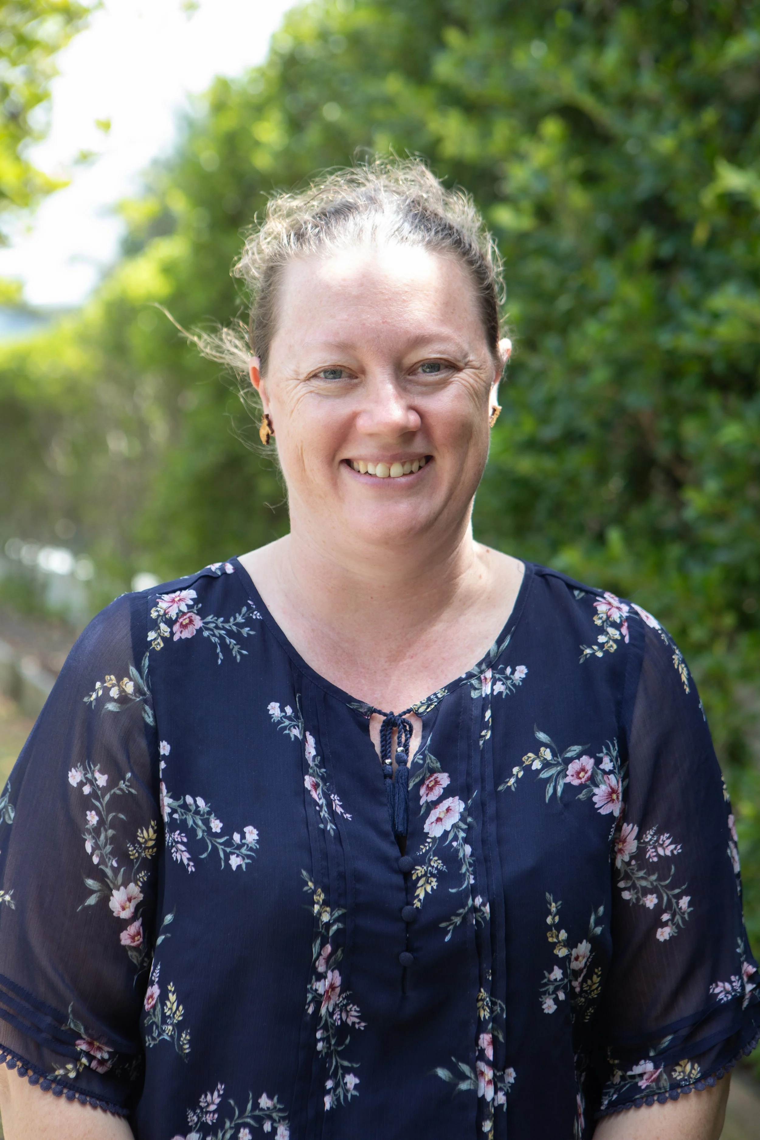 Smiling woman with light brown hair tied back, wearing a navy blue floral blouse, standing outdoors with green foliage in the background.