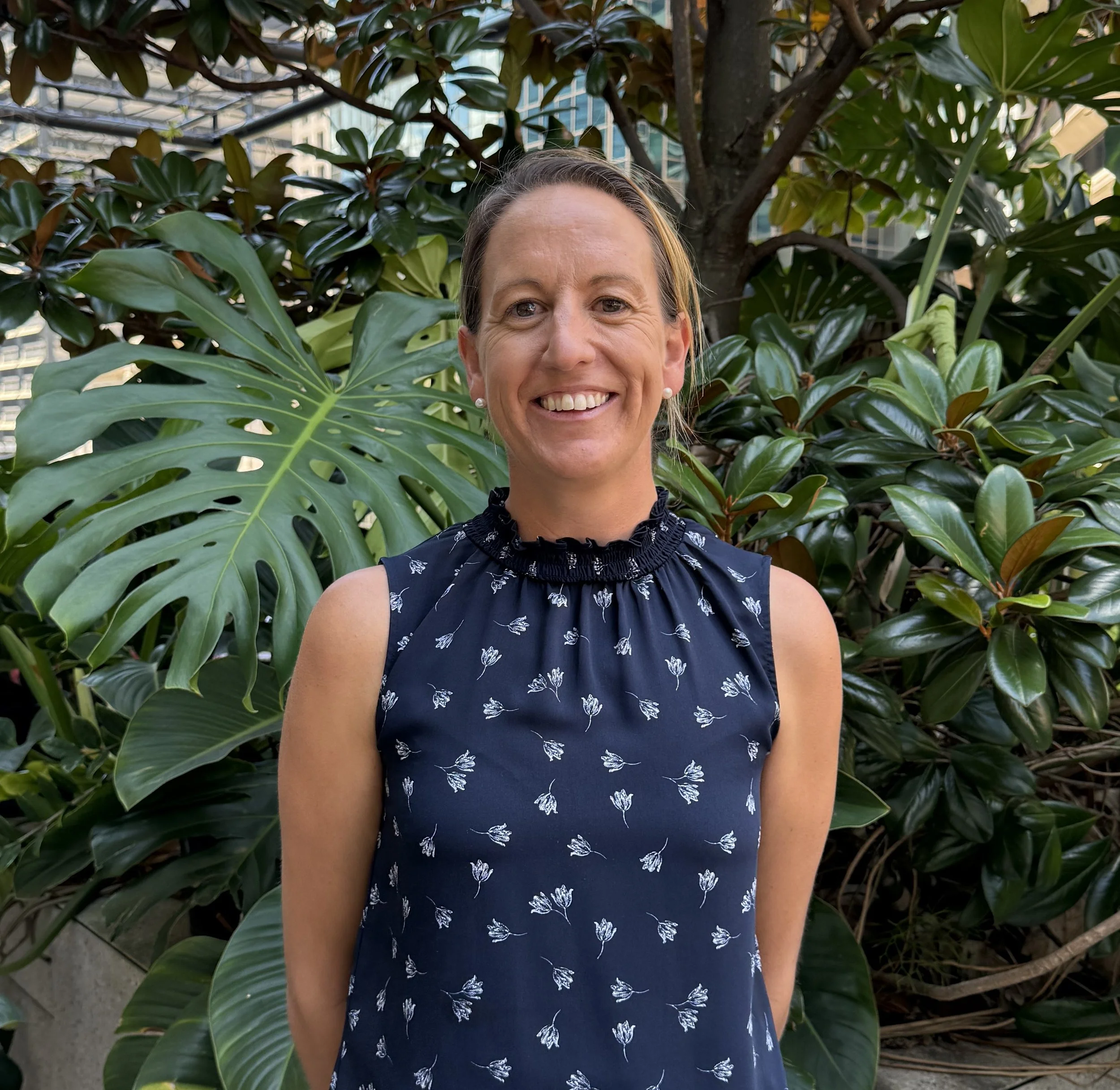 A woman with light skin, wearing a sleeveless navy dress with white floral pattern and ruffled high neckline, standing outdoors in front of large green tropical leaves and trees.