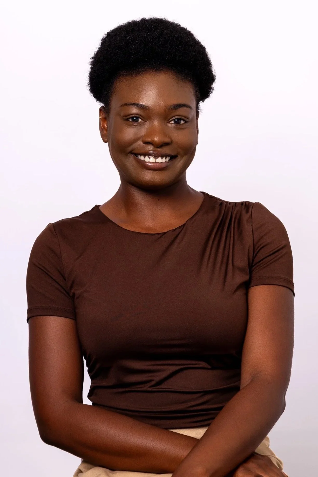 A smiling Black woman with short curly hair, wearing a brown short-sleeved top, standing against a plain white background.