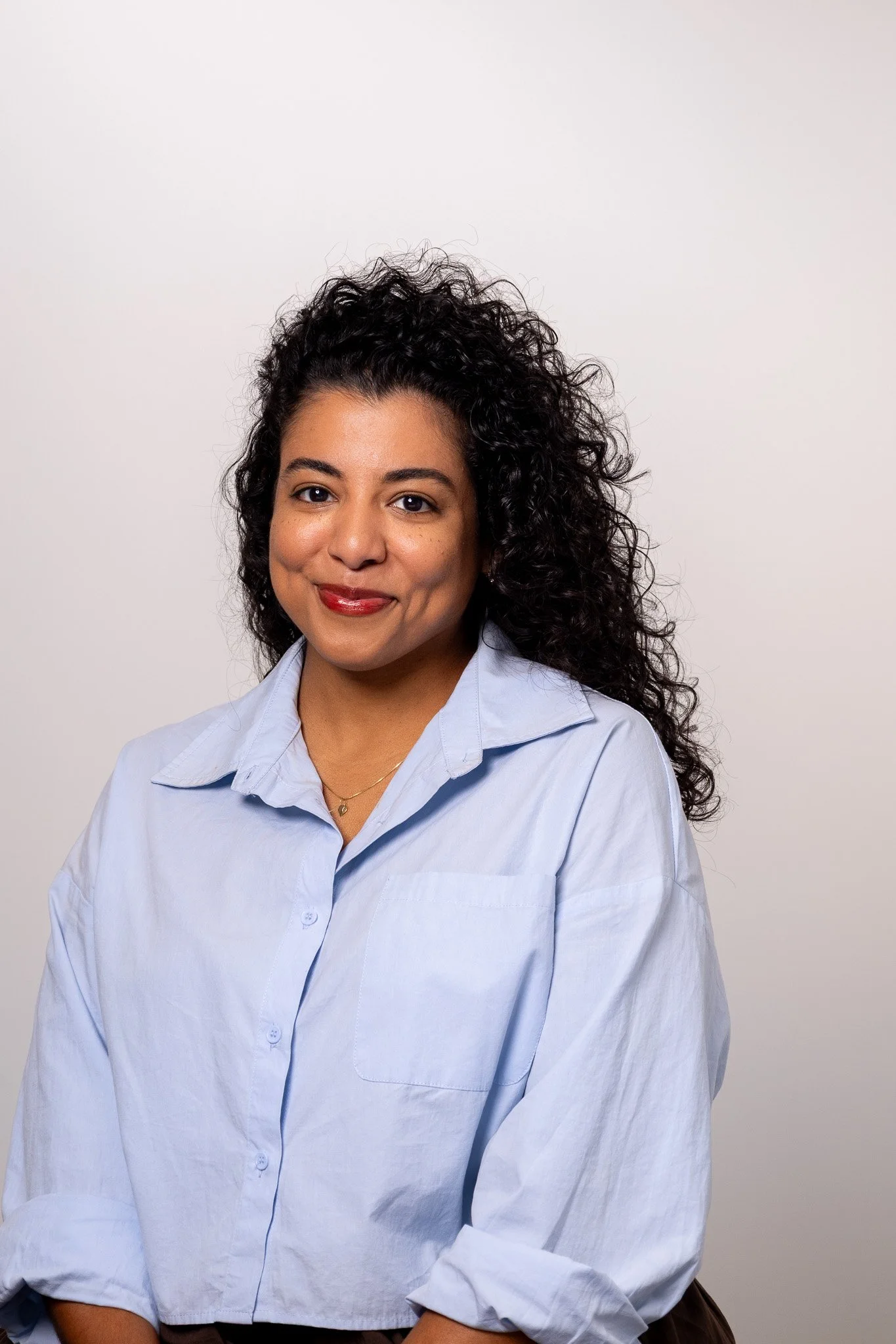 A woman with curly black hair smiling, wearing a light blue button-up shirt against a neutral background.