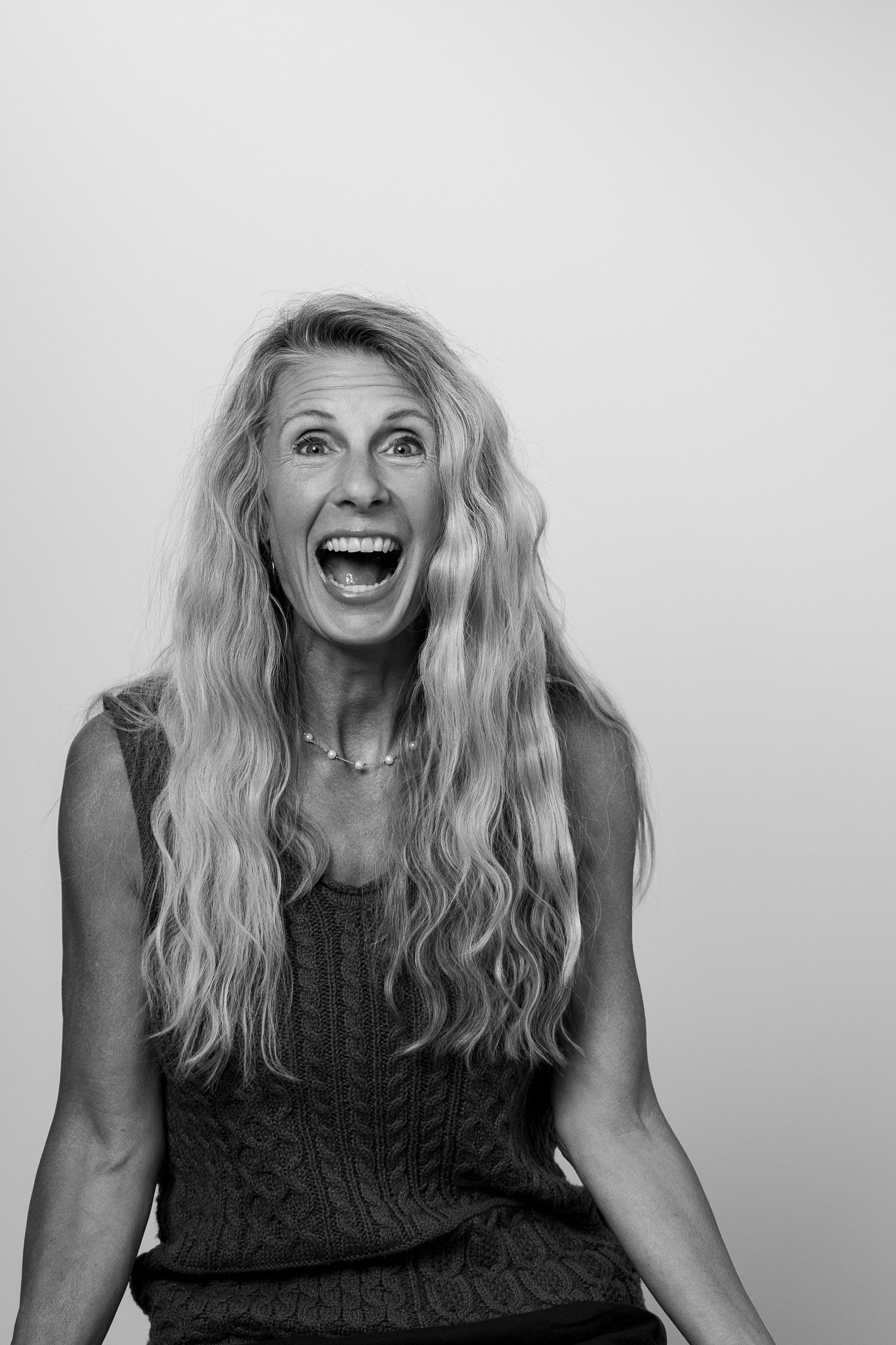 Black and white photo of a woman with long, wavy hair, smiling widely with her mouth open, wearing a sleeveless knit top and a pearl necklace, against a plain light background.