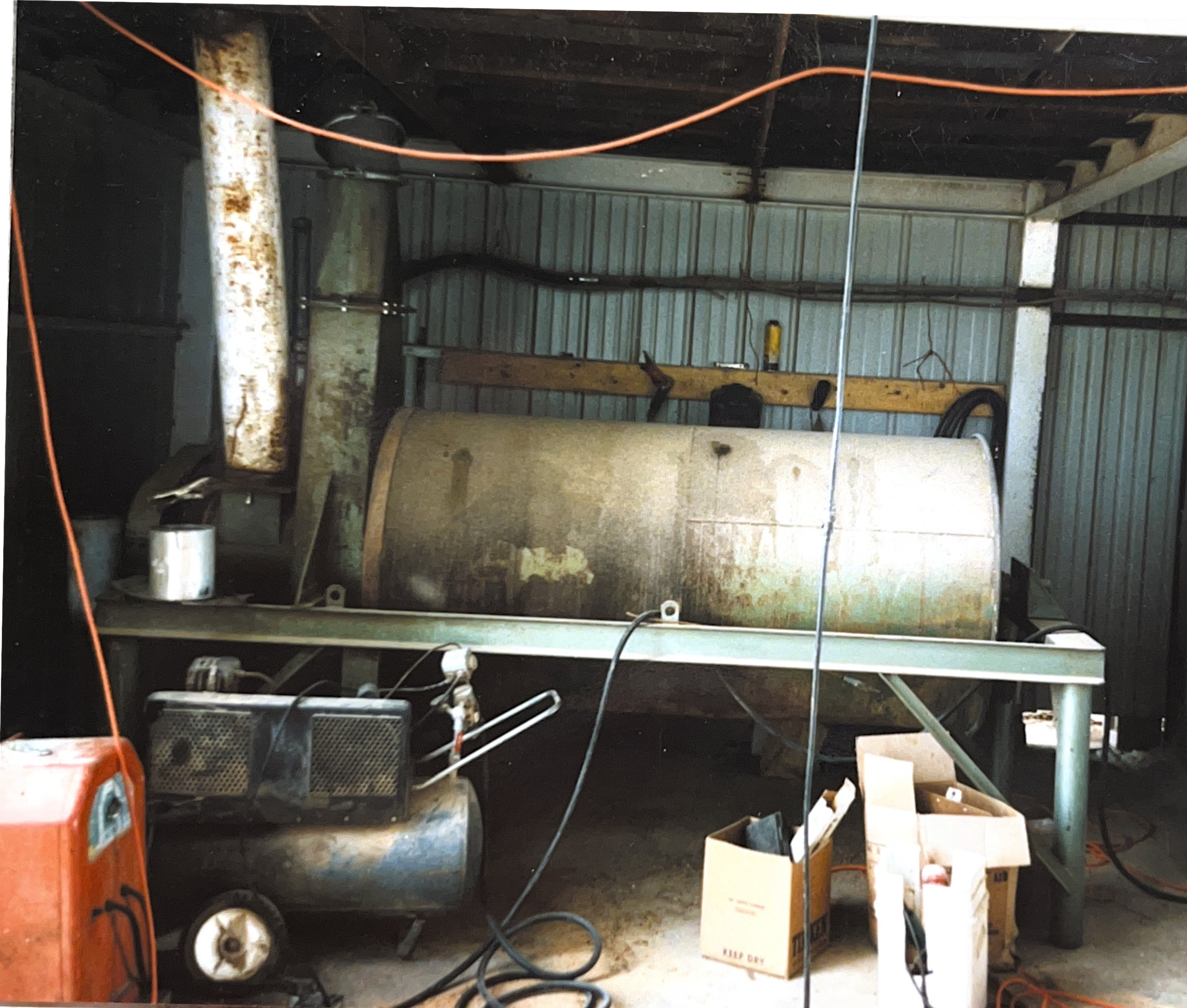 A large rusty cylindrical tank on a metal frame inside a shed with metal walls. Various tools, wires, and boxes are scattered around.