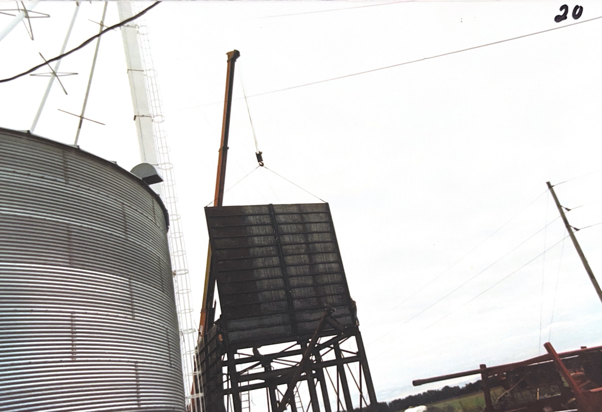 A grain silo, a crane lifting a large black container or elevator, and utility poles with wires against an overcast sky.