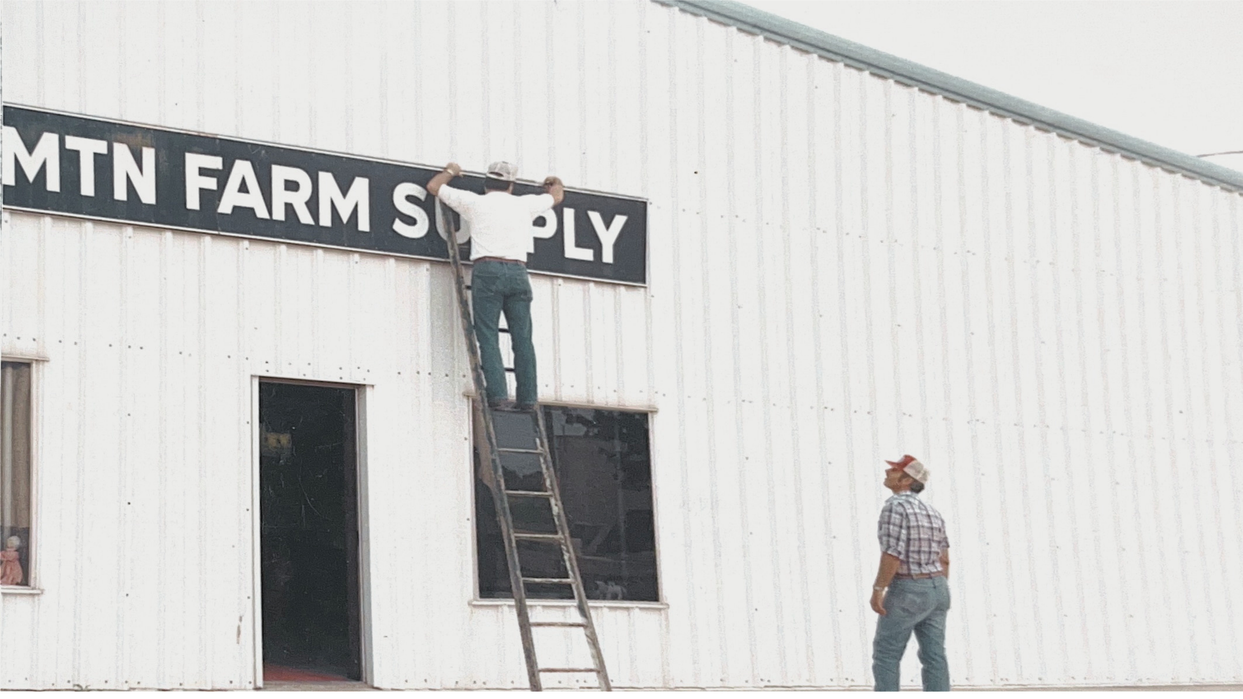 Two people are installing a sign on an exterior white metal building. One person is standing on a tall wooden ladder, holding a pole with the sign, while the other person looks on from the ground.