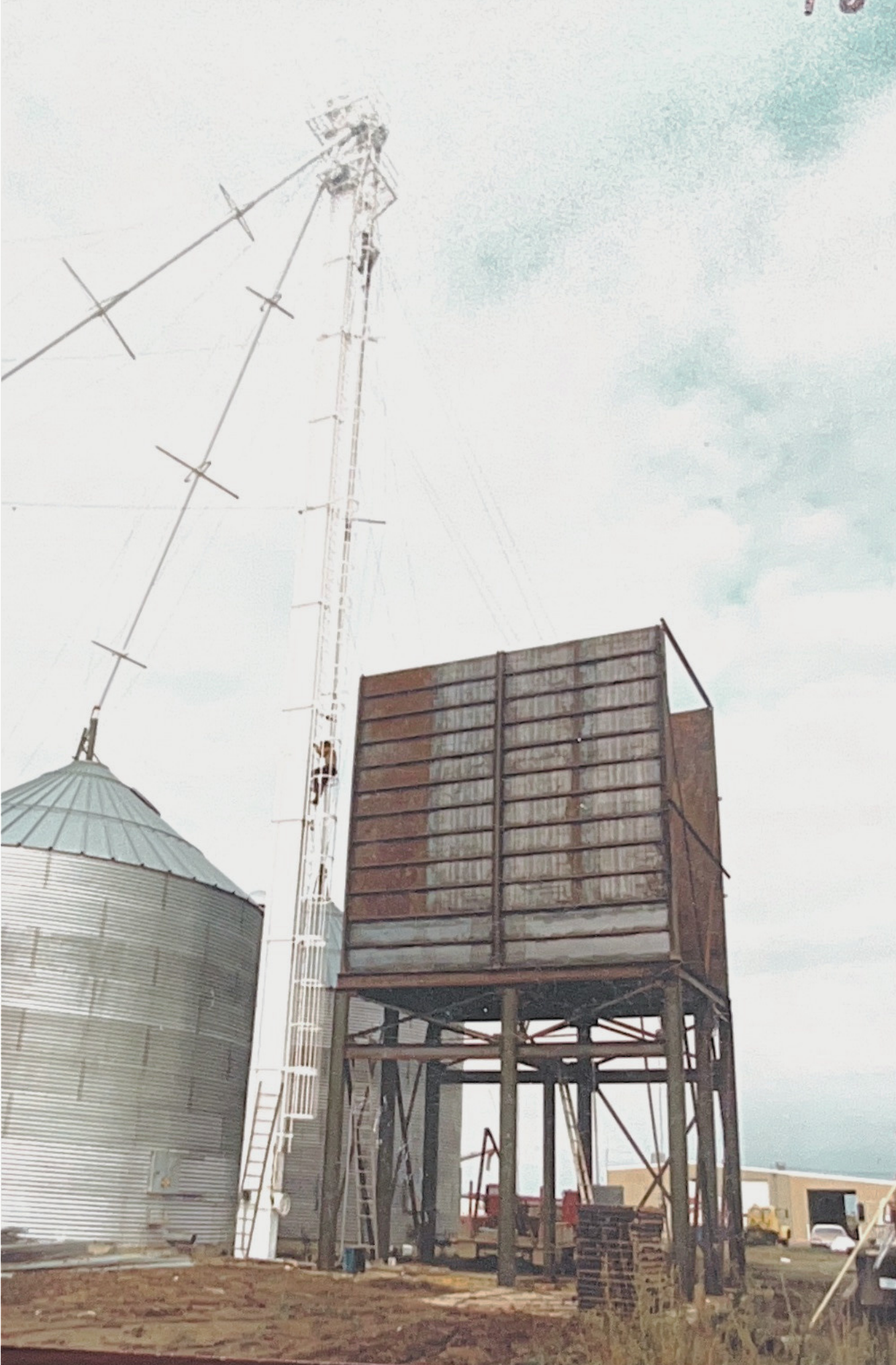An outdoor scene with a metal grain silo on the left, a tall communication tower with antennas to the left of a wooden elevated water tank, which stands on metal legs. The sky is cloudy.