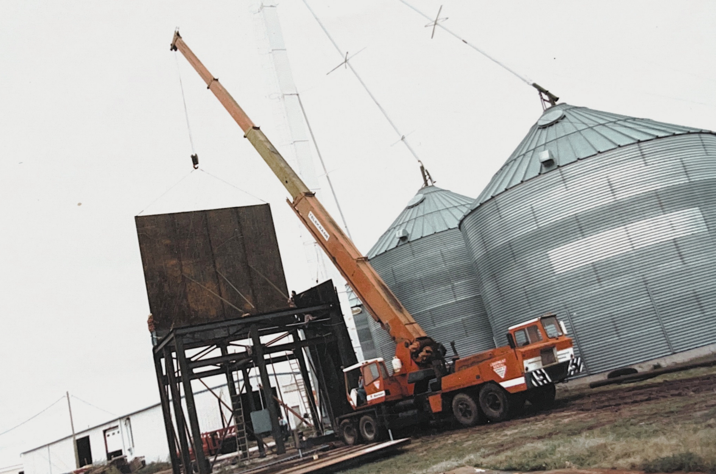 A construction crane lifting a large black metal object next to a silvery grain silo on a farm.