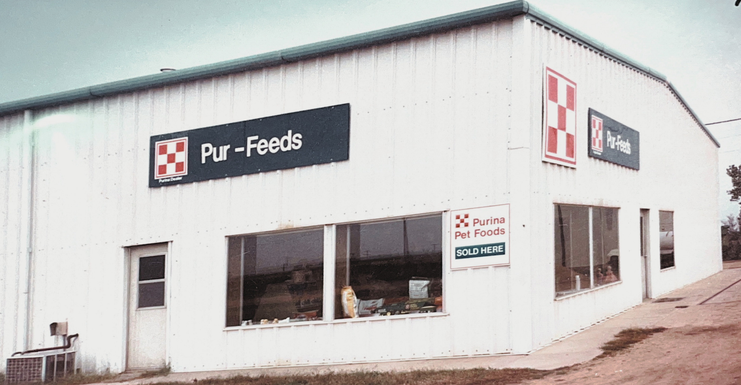 White commercial building with signs for Pur-Feeds and Purina Pet Foods, located on a dirt lot with a dirt pathway in front.