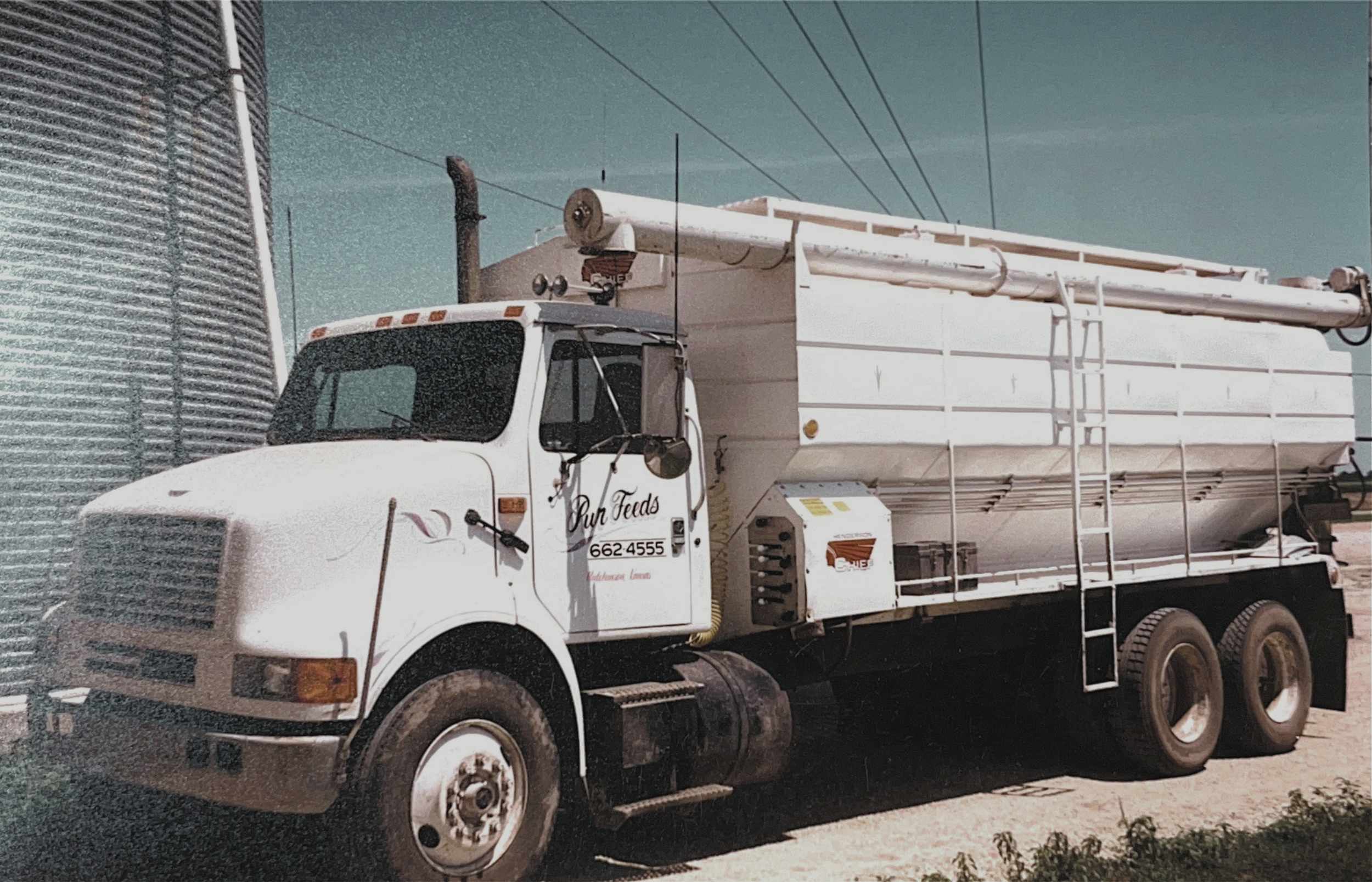 White milk delivery truck with the text 'Ryn Feeds' on the door, parked outdoors near a large silver grain silo, with clear blue sky overhead.