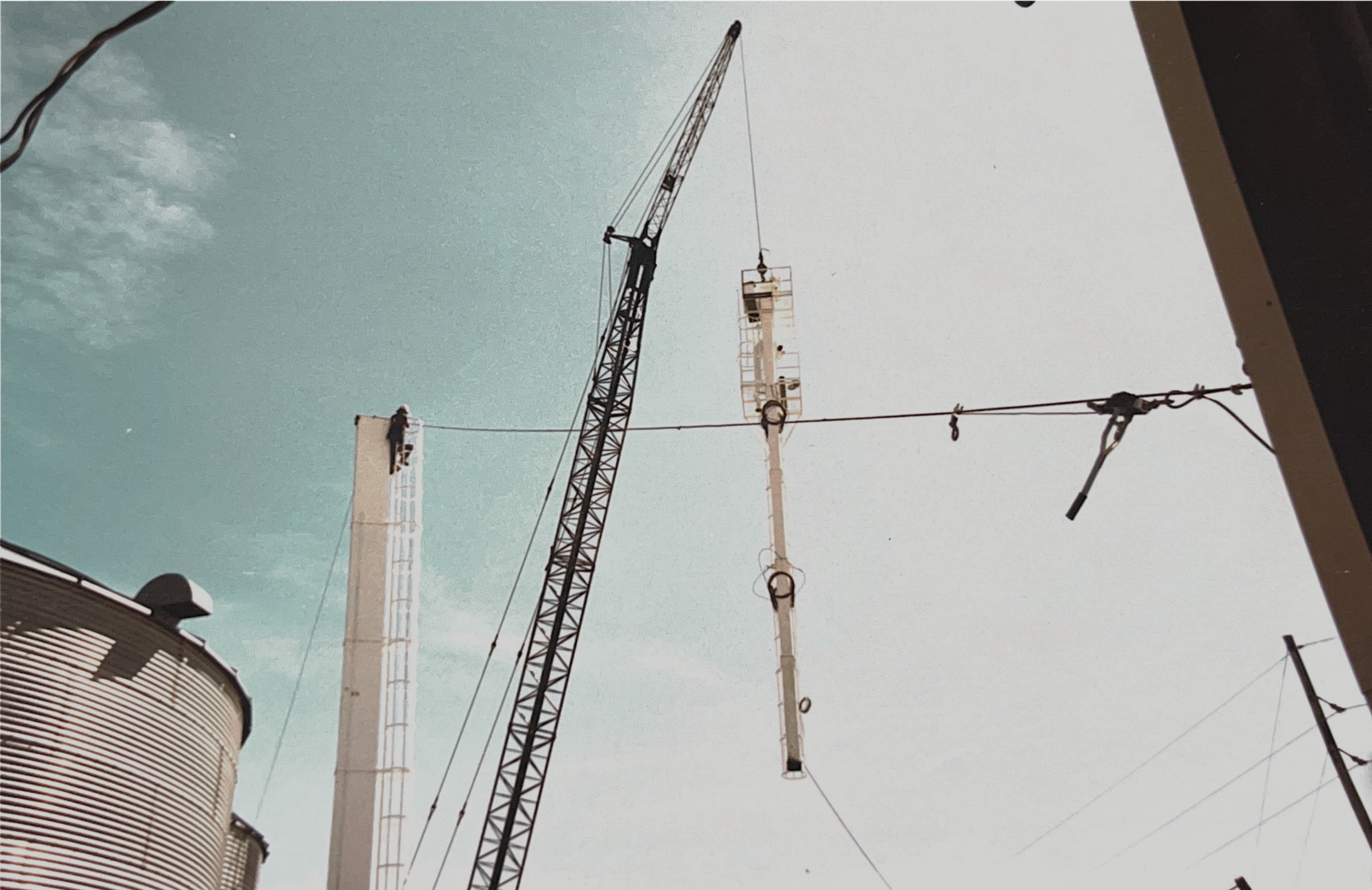Construction crane lifting a large piece of equipment against a partly cloudy sky, with power lines in the foreground and industrial buildings on the side.