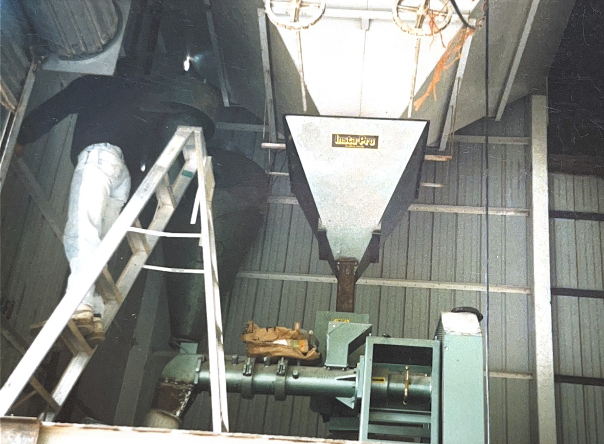 Industrial machine with a funnel labeled 'Insta Pro', ladder, and a person working on the machinery inside a high-ceiling industrial building.