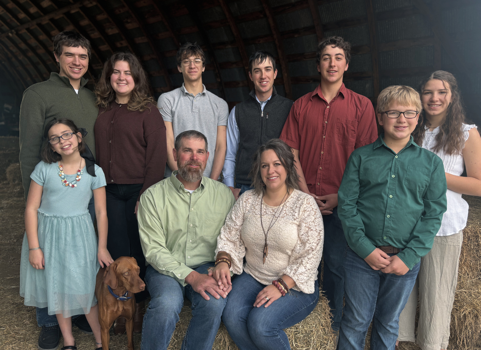 A family of twelve people and a dog posing inside a barn with hay on the floor and a wooden ceiling.