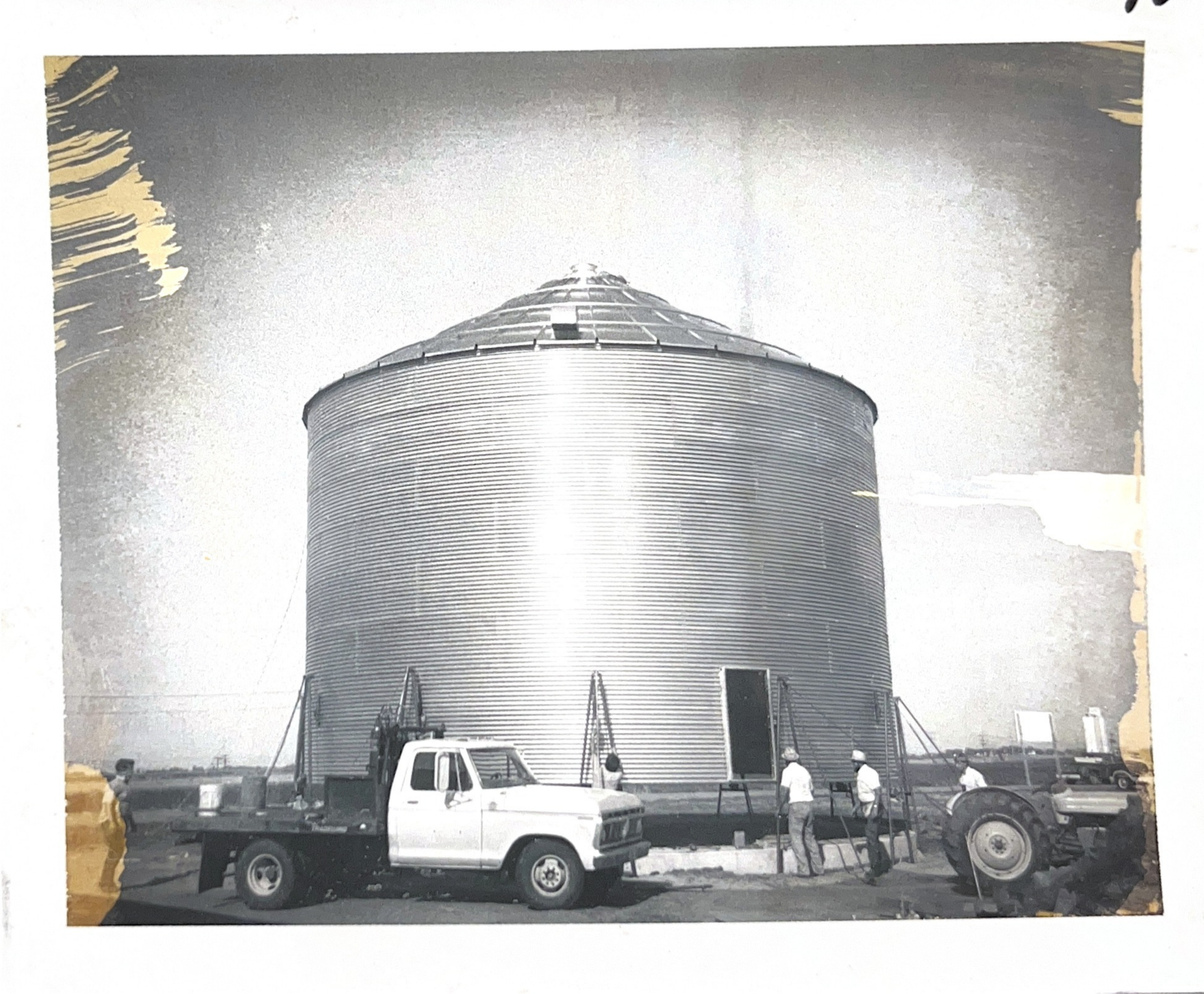 Black and white photo of a large cylindrical grain silo with a conical roof, surrounded by construction vehicles and workers.