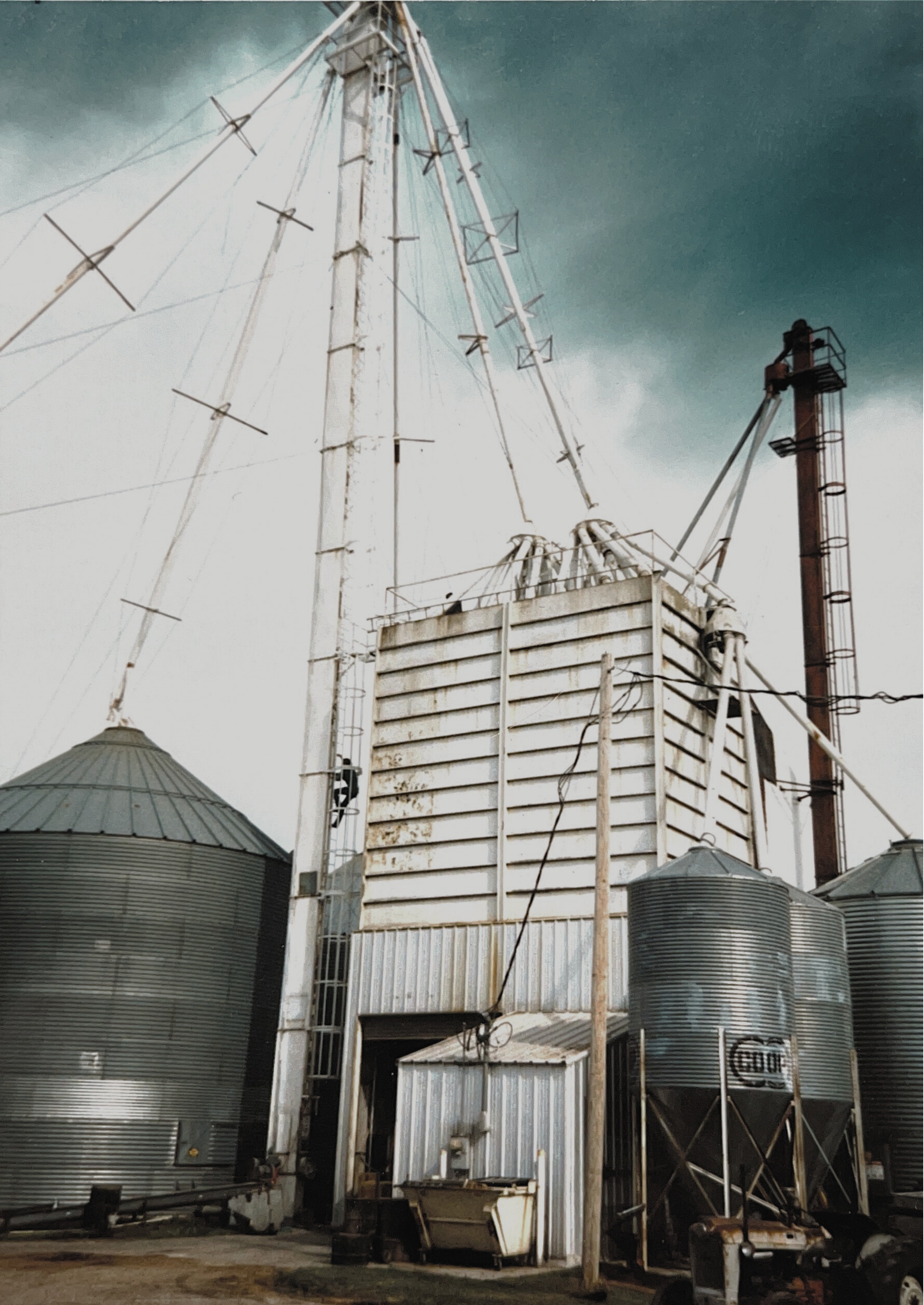 Farm storage silos and a grain dryer with a tall metal structure, set against a cloudy sky.