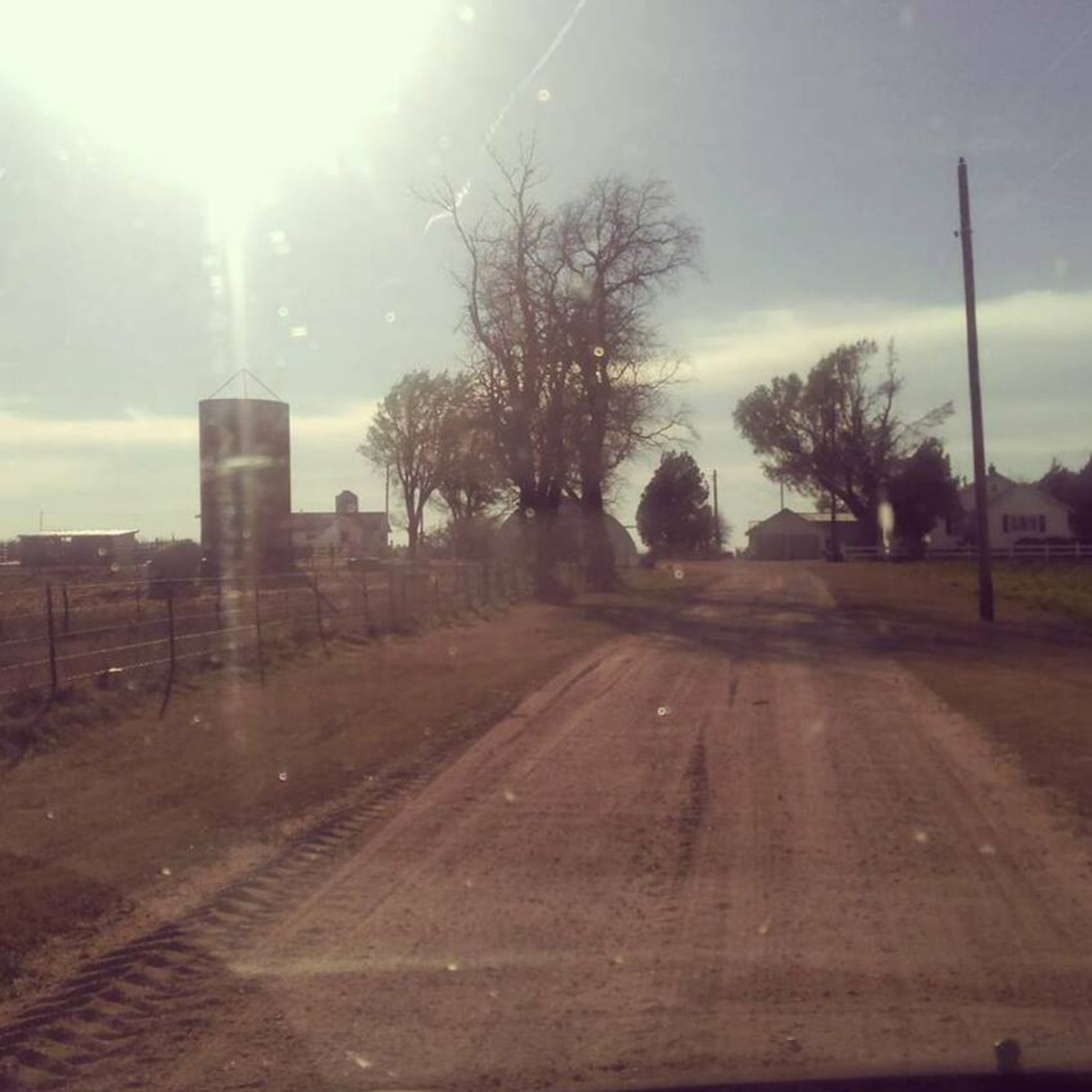A dirt road through a rural farm area with trees, a silo, and a few houses under a partly cloudy sky.