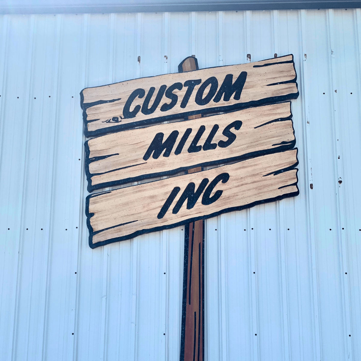 Wooden sign with black painted text reading "Custom Mills Inc" attached to a metal wall.