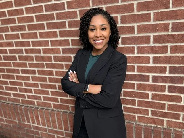 A photo of Dr. Wilson - a Black and Chicana woman with curly black hair, wearing a dark blazer over a green top, standing with arms crossed in front of a brick wall, smiling.