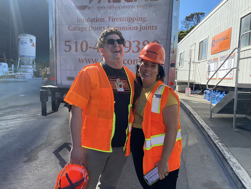 Picture of Sam Moss, Executive Director of Mission Housing, on left in orange safety vest with hat in hand and Youth Art Exchange Executive Director Raffaella Falchi Macias in orange safety vest with hat on smiling at construction site.