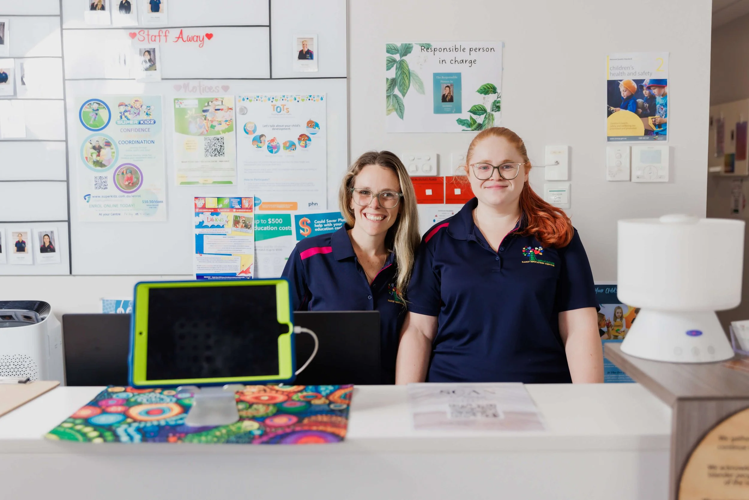 Two smiling female childcare workers in uniform standing behind a desk with educational posters hung in the background.