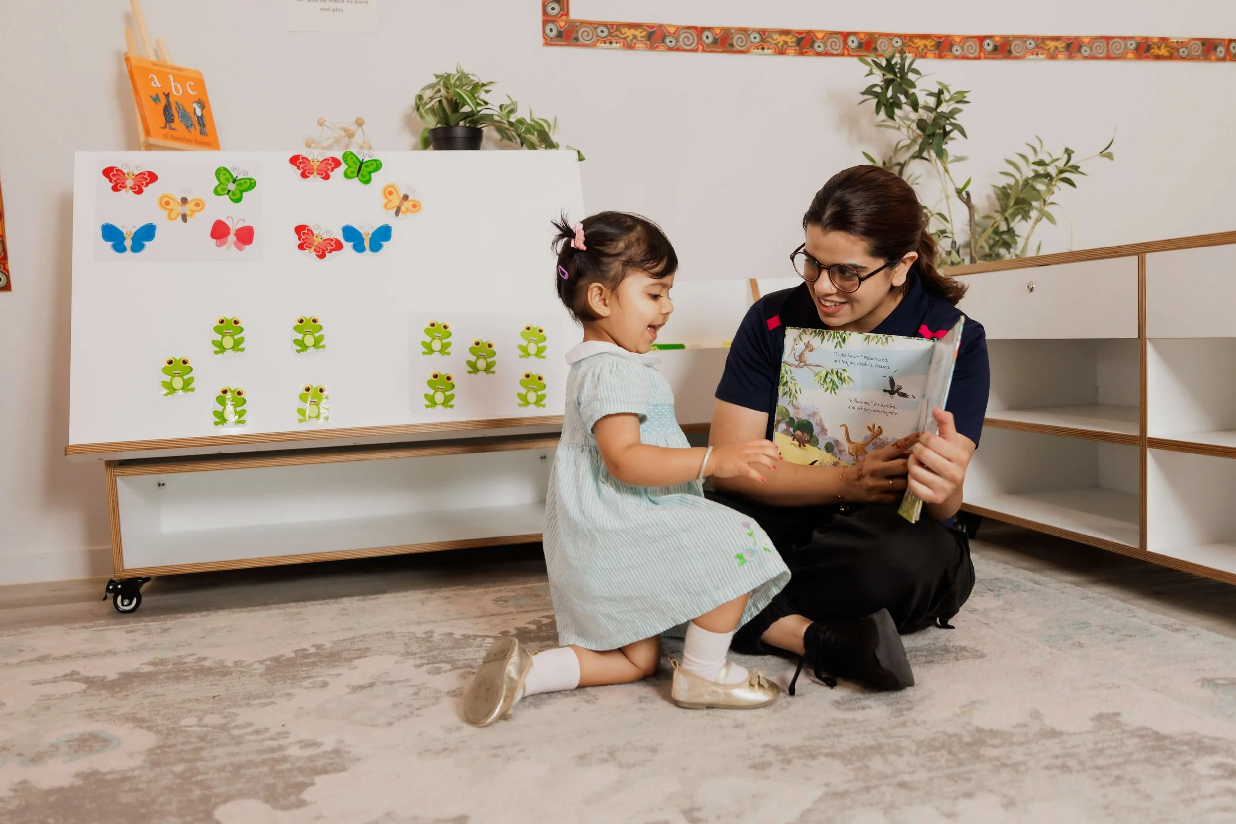 A childcare worker sitting on carpet next to a girl and reading a picture book to her.