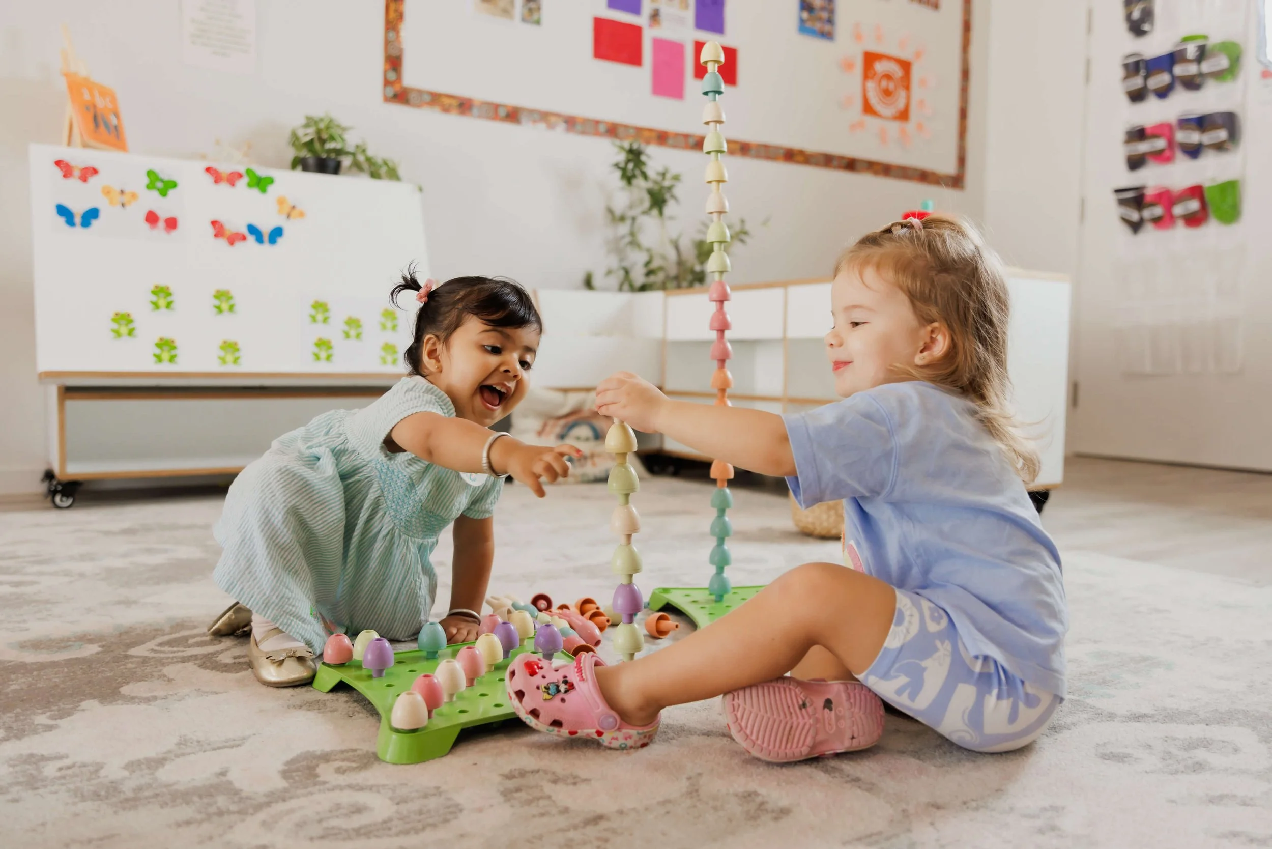Two smiling children play will coloured toys to create a tower in a childcare centre.