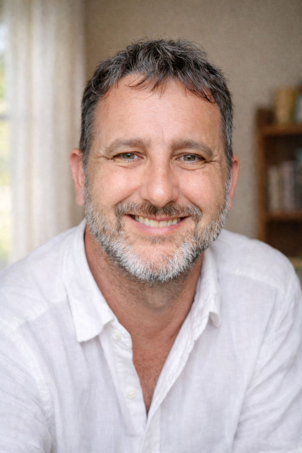 A smiling middle-aged man with gray hair and beard, wearing a white shirt, indoors with a bookshelf in the background.