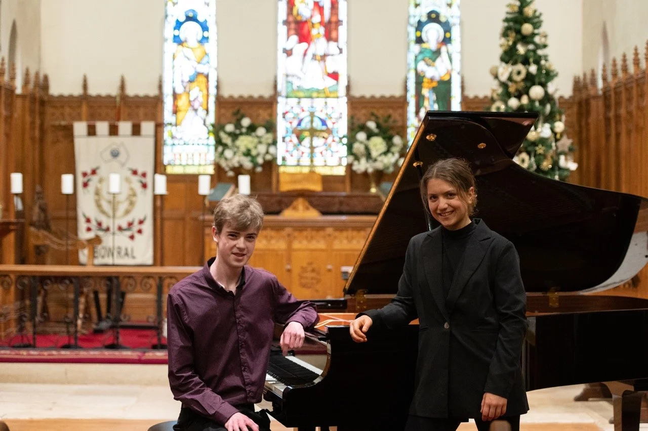 A young man and a young woman in formal attire posing with a grand piano in a church or hall with stained glass windows, Christmas tree, and decorations.