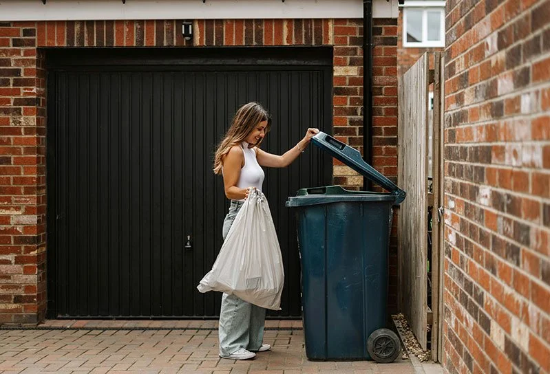 Young woman throwing trash into a blue garbage bin outside a brick house.