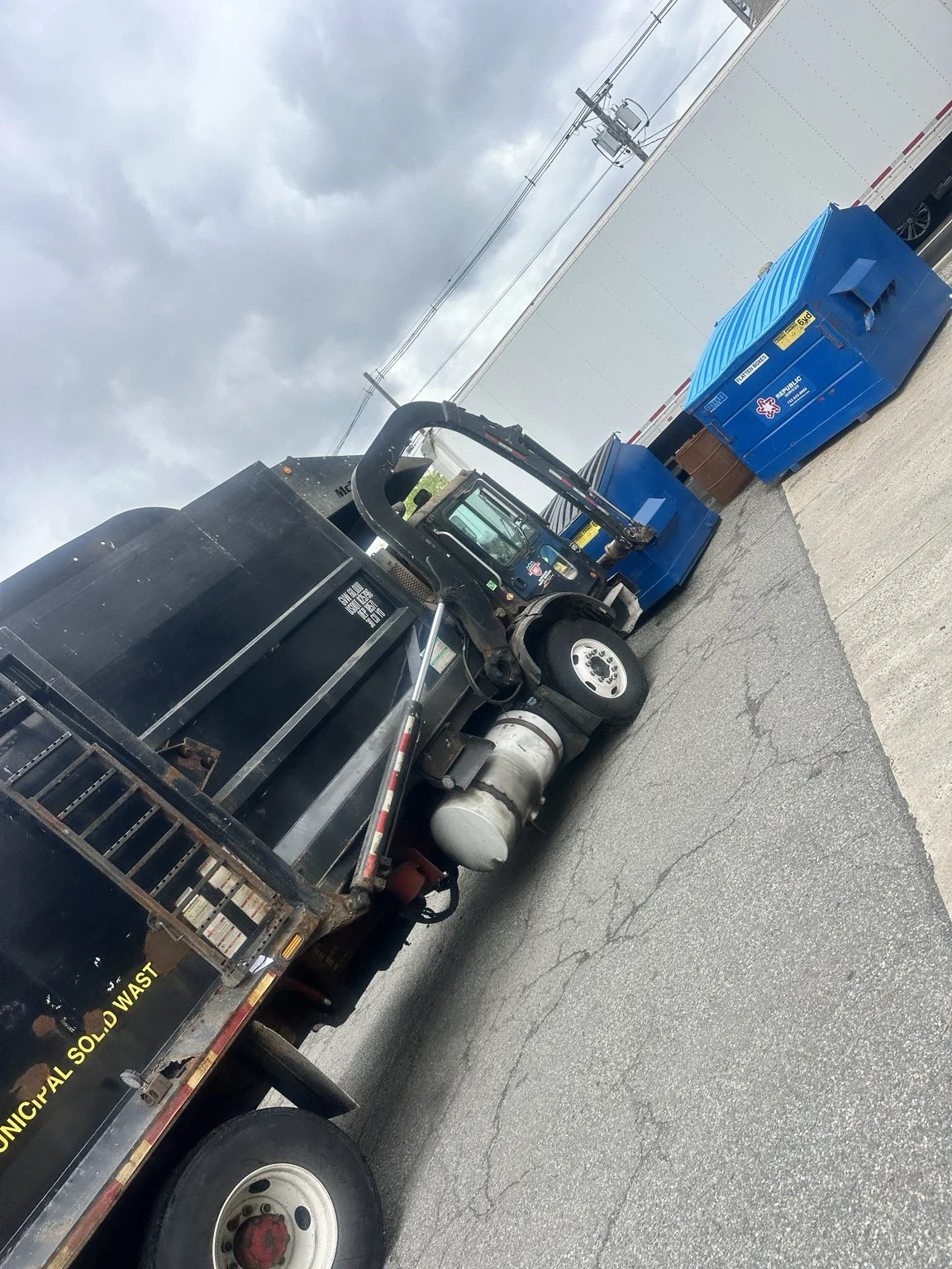 An industrial street scene showing a black garbage truck with a hydraulic arm, blue recycling bins, and a utility pole with power lines against a cloudy sky.