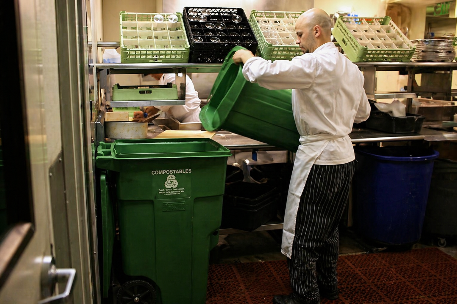Chef placing a green compostable bin into a larger green compost bin in a restaurant kitchen with other bins and shelves around.