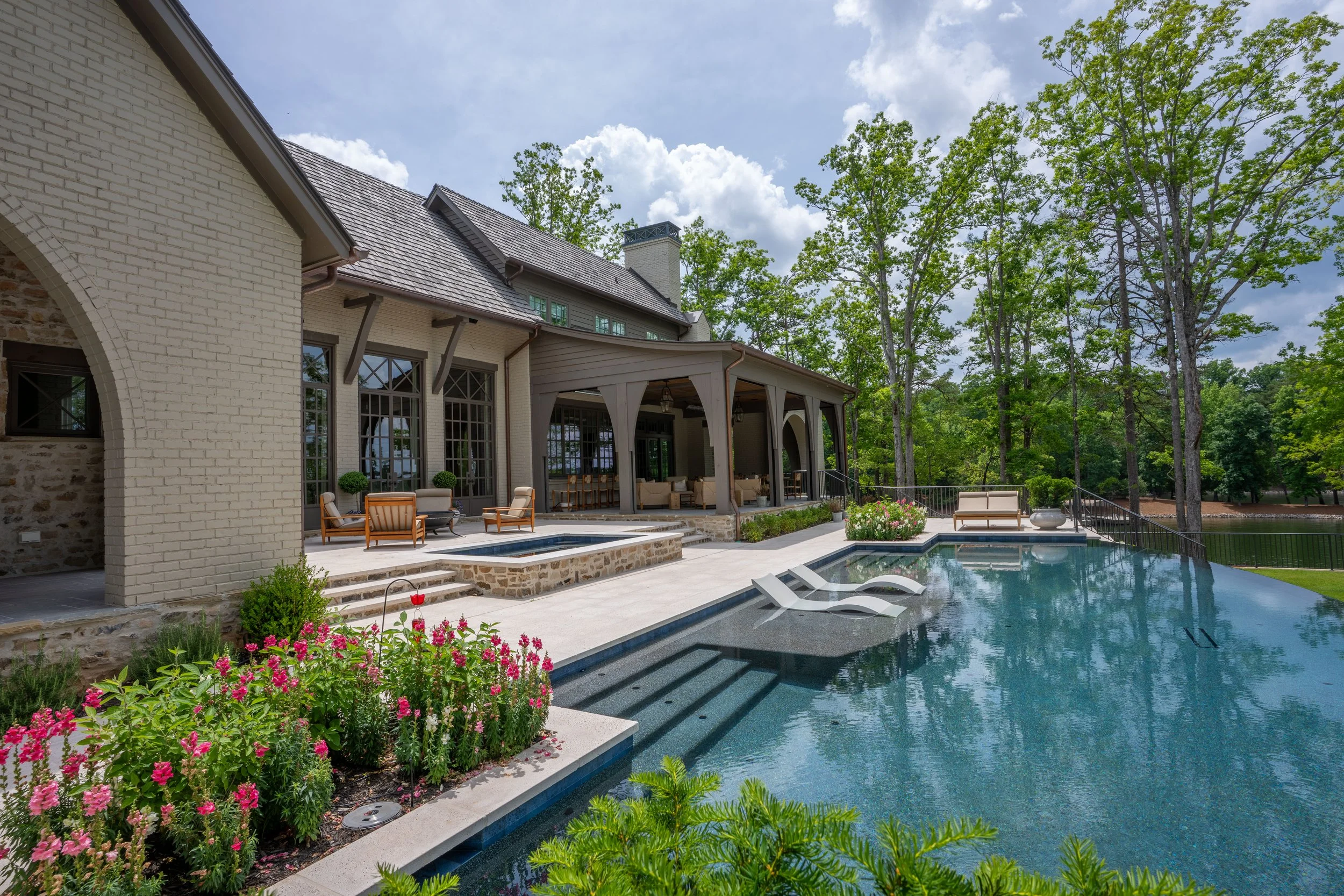 Luxury backyard with swimming pool, lounge chairs, patio with seating, and house with large windows and trees in the background.