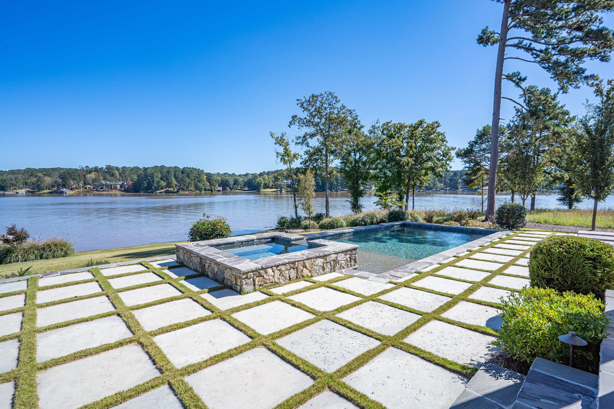 View of a backyard with a pool and hot tub overlooking a lake, with trees and houses in the distance, under a clear blue sky.